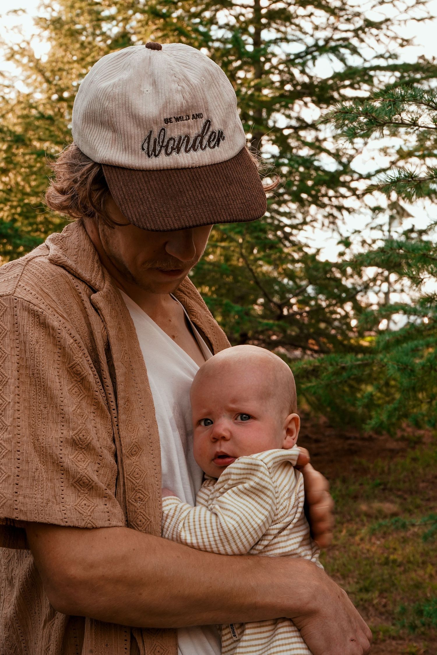 A person wearing a corduroy cap tenderly holds a baby in striped clothing outdoors.