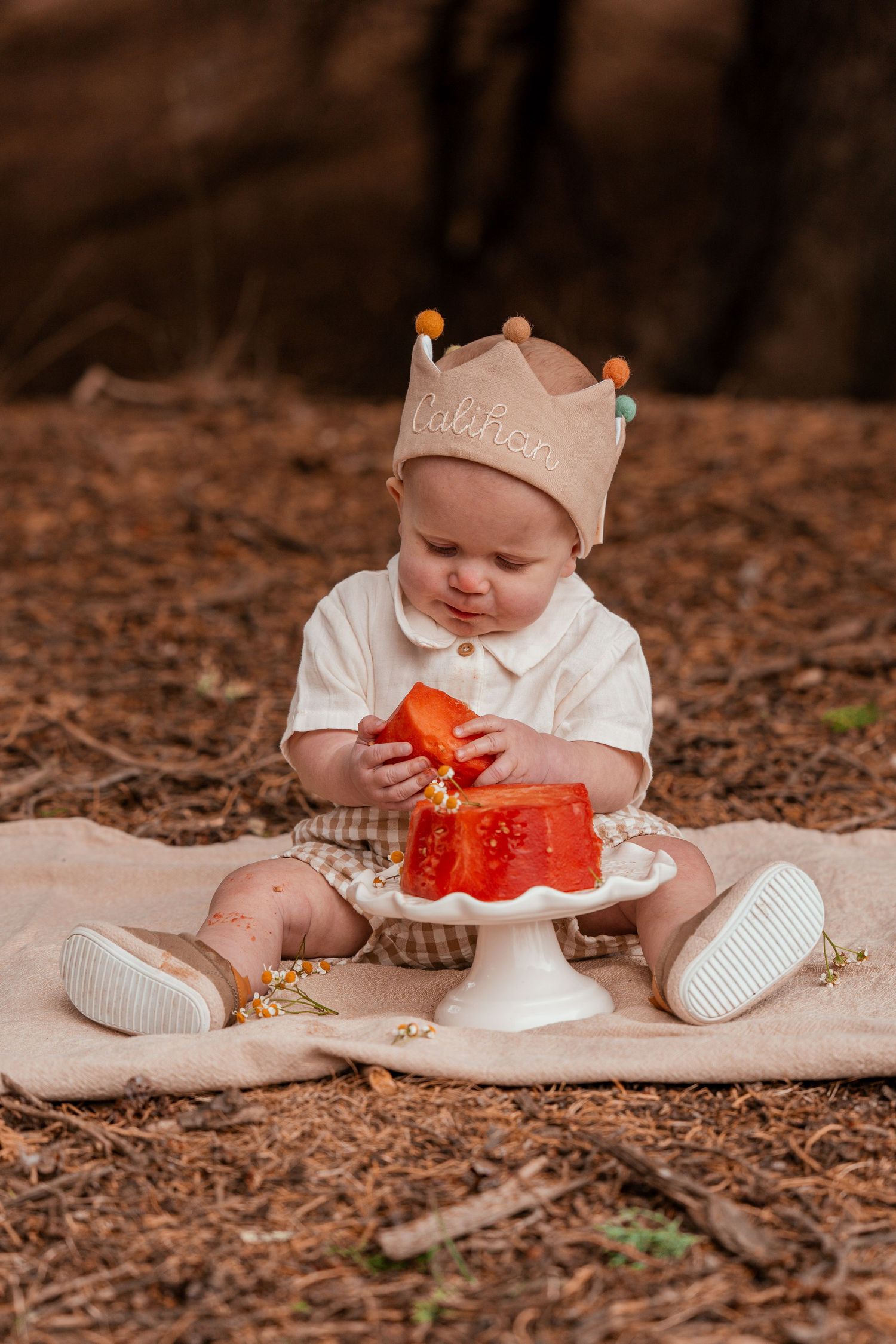 Baby in white outfit and crown hat sits on blanket with orange jello cake on pedestal plate in outdoor setting.