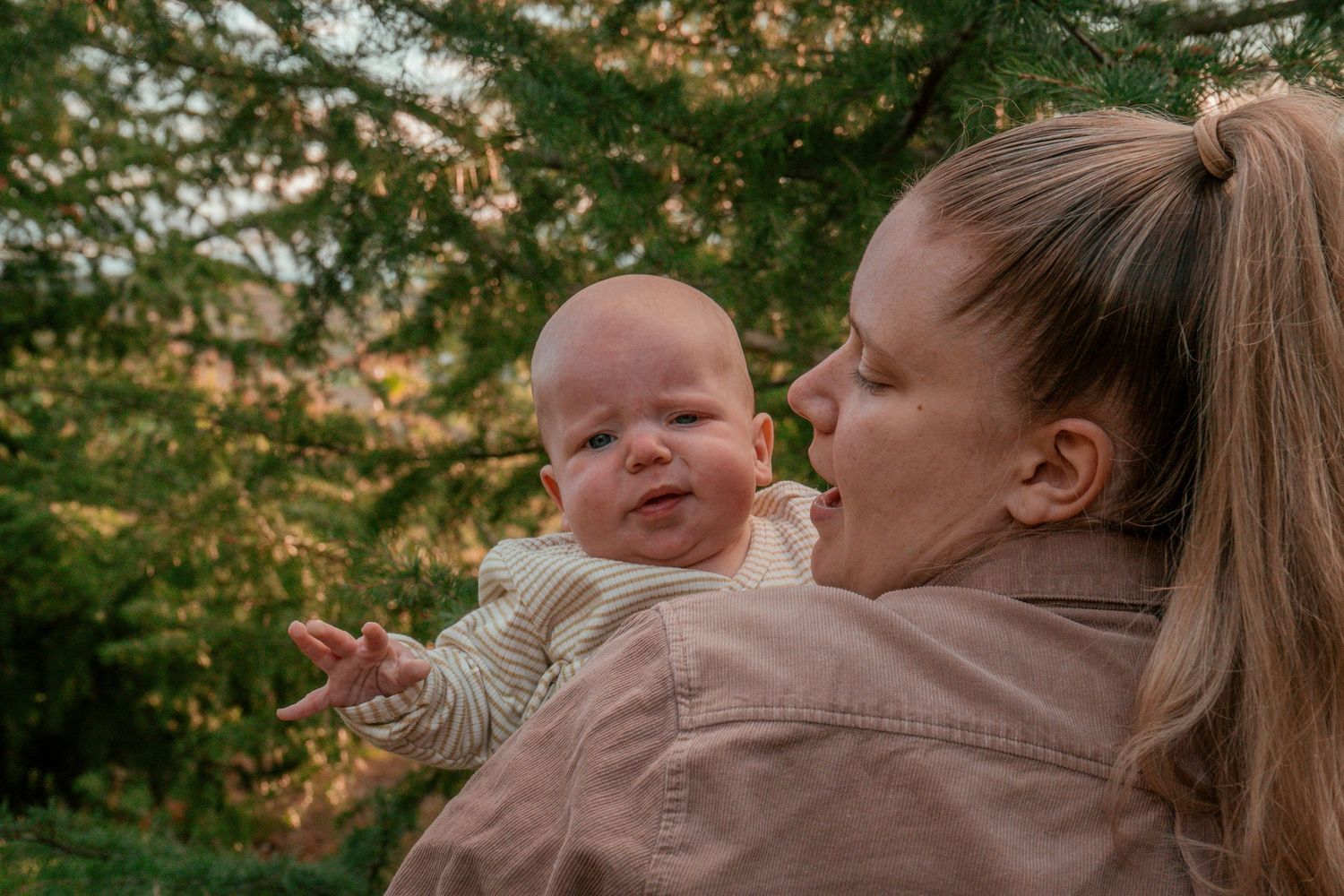 Sweet candid moment between parent and infant outdoors near pine trees.