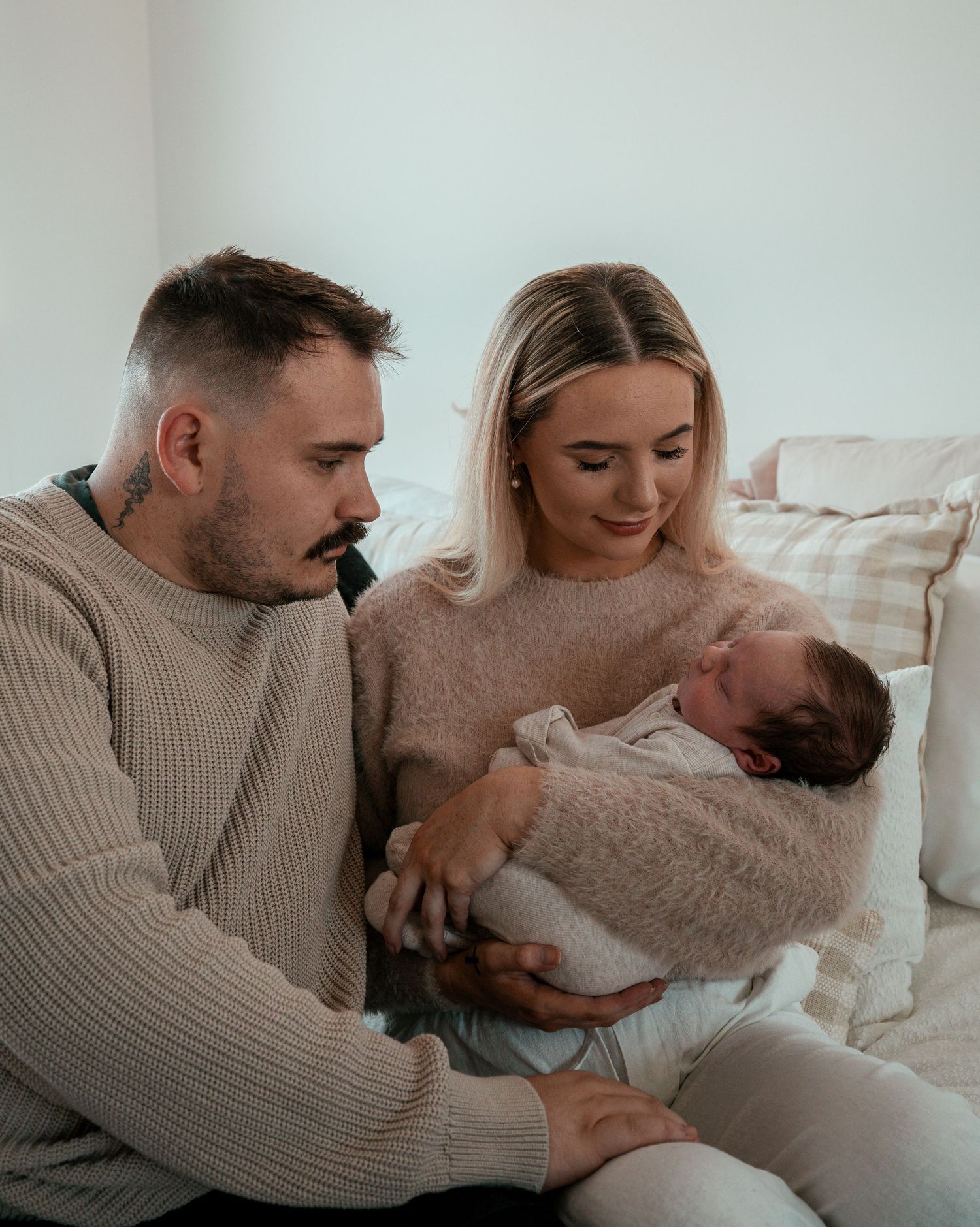 Parents in matching beige sweaters lovingly gaze at their newborn baby wrapped in a cozy blanket.