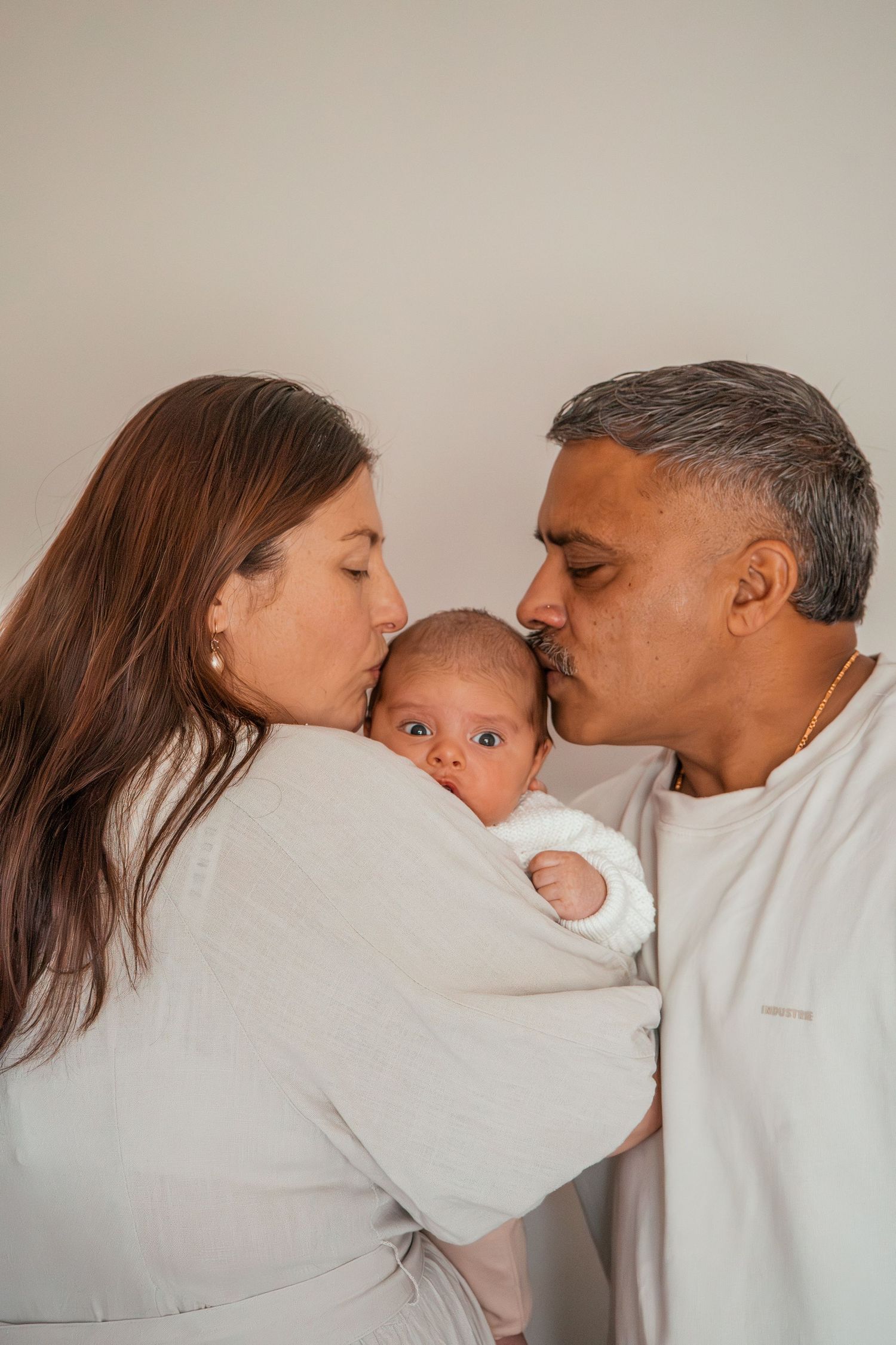 Family sharing tender kisses with their baby in a series of intimate portrait shots against a neutral wall.