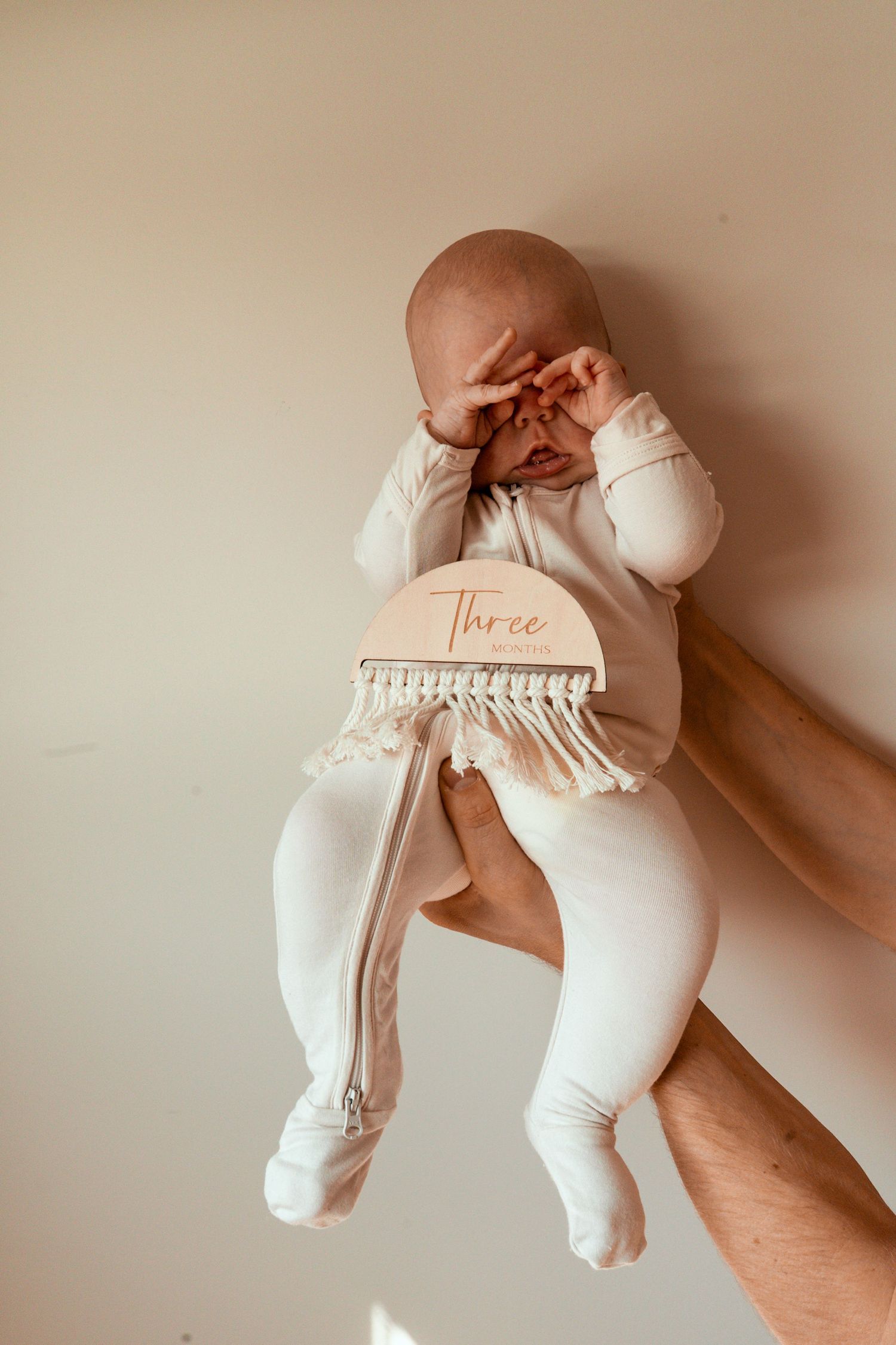 Baby in white outfit holding a decorative milestone sign with tassels that says 'Three'.