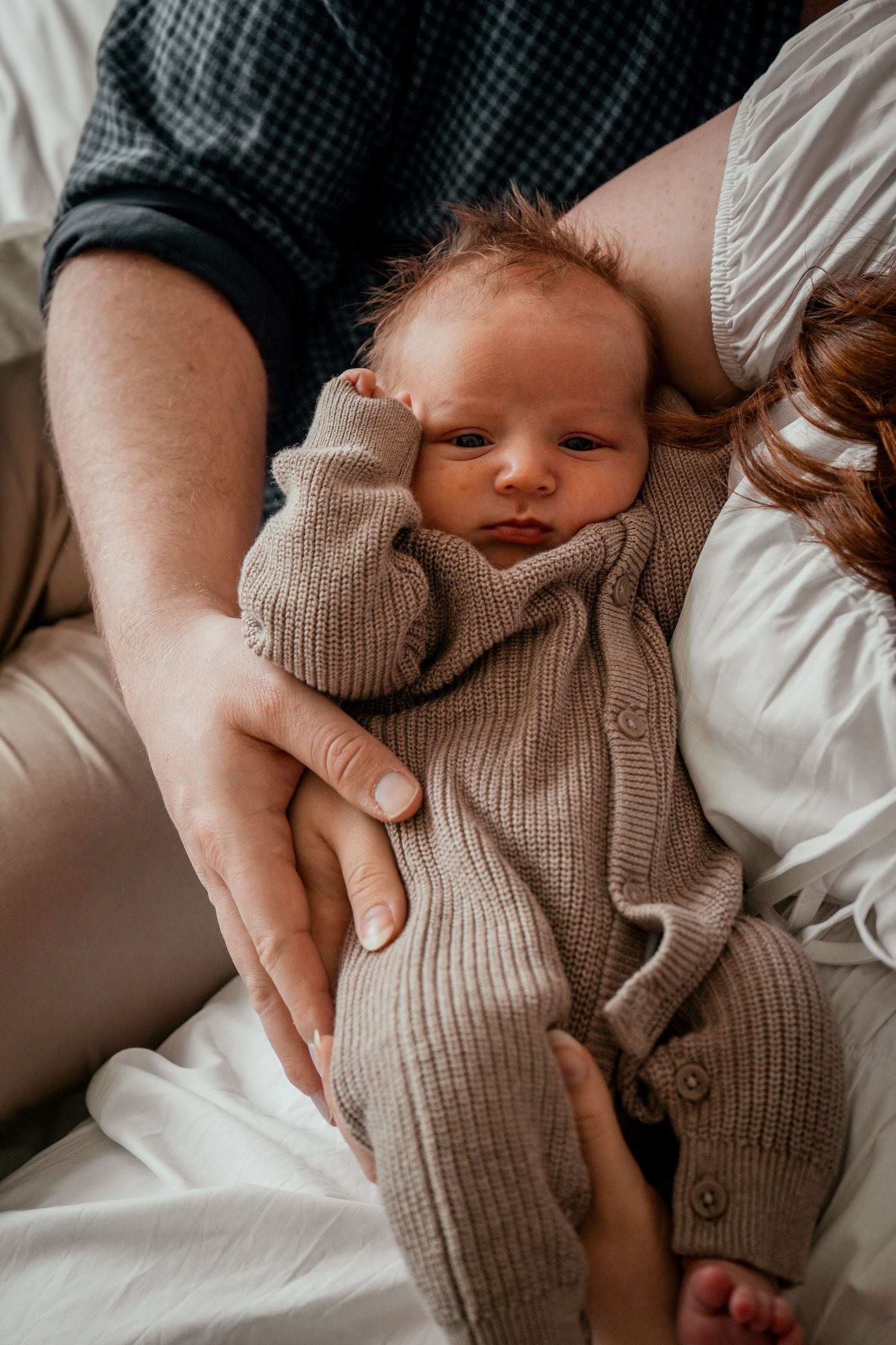 Baby wearing cozy knit sweater lies on white bedding while being cradled by gentle hands.