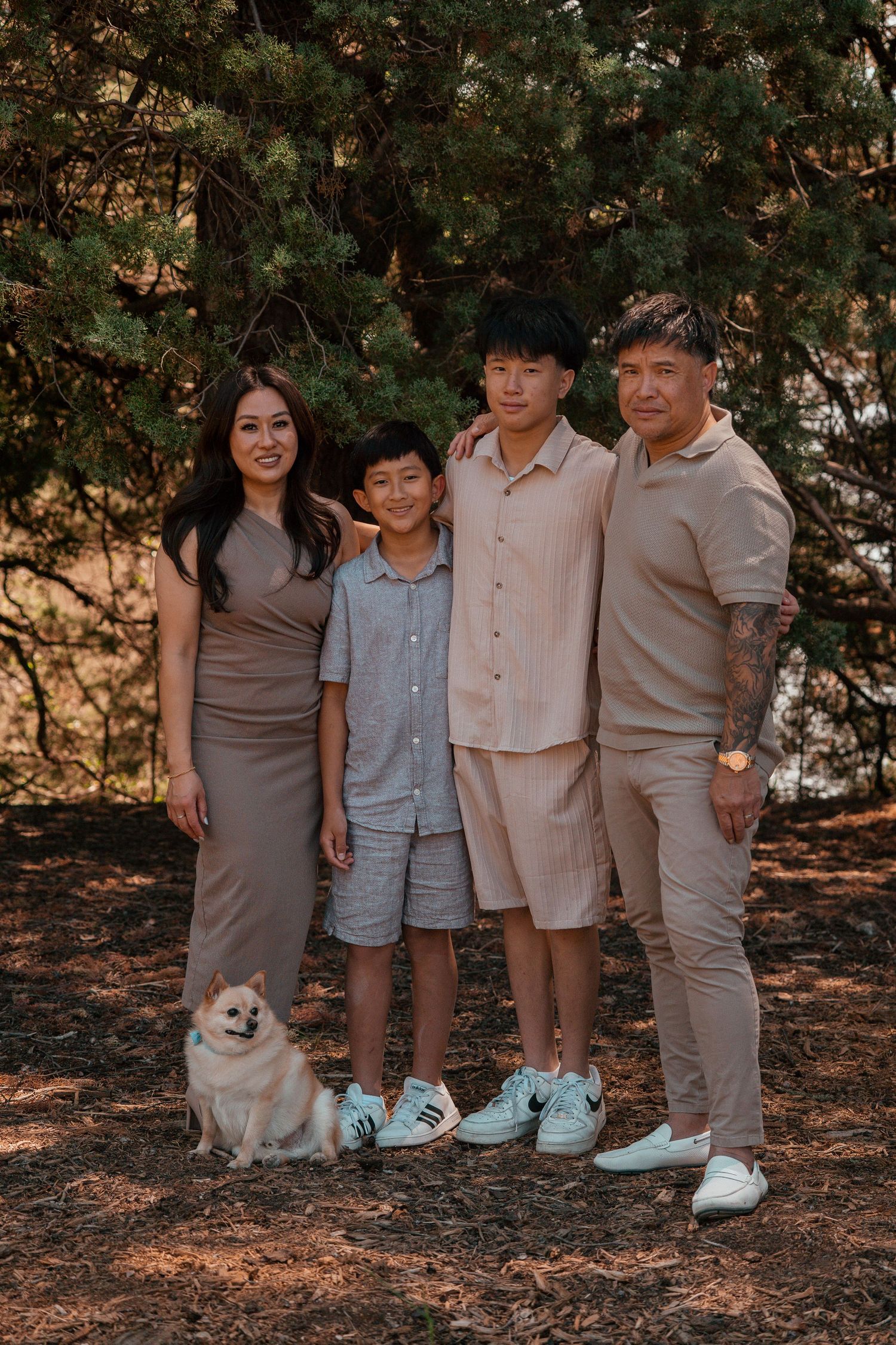 Family in neutral-toned clothing poses with their white dog on pine needle covered ground.
