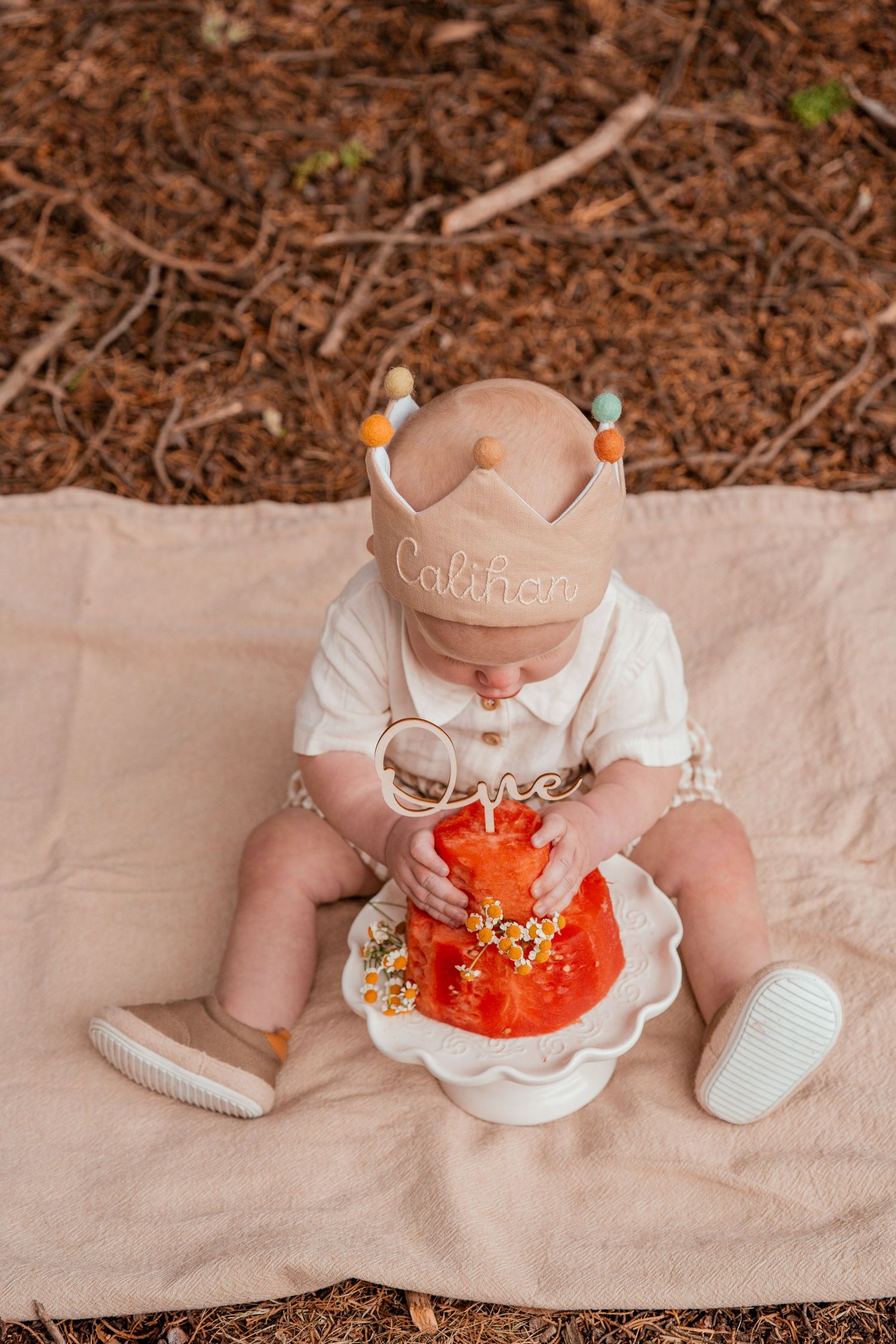 A baby wearing white sits on a beige blanket while holding and playing with a bright orange plush toy.