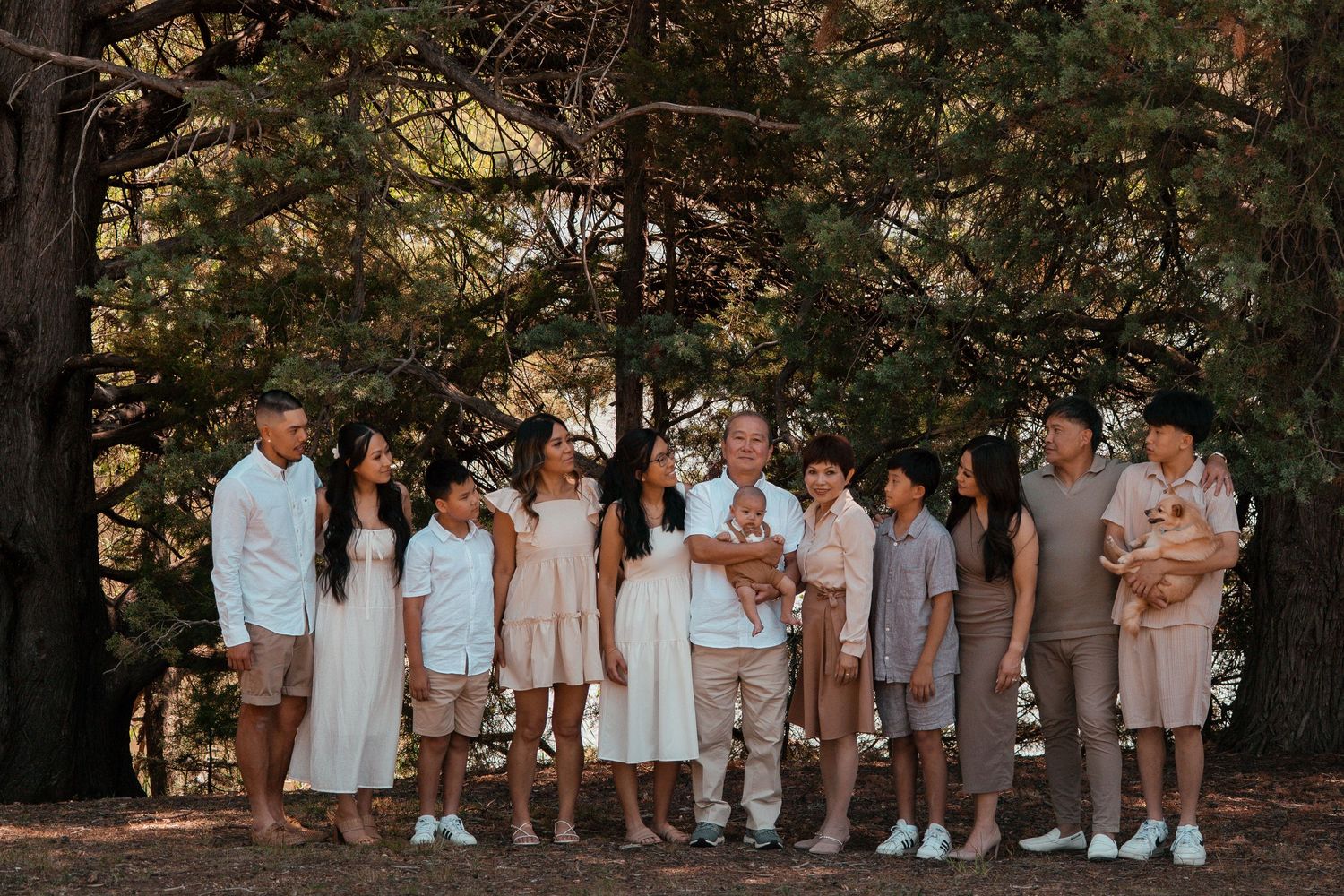 A group in coordinated neutral and white outfits poses together in a wooded outdoor setting during a photo session.