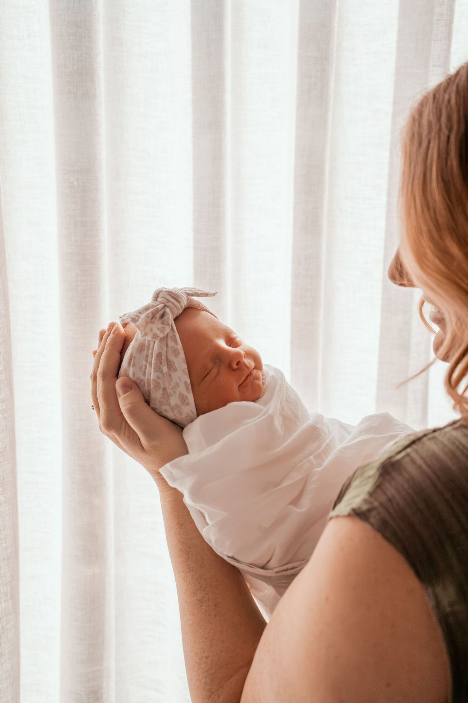 A baby in white clothing is being held up against a bright window with white curtains in the background.