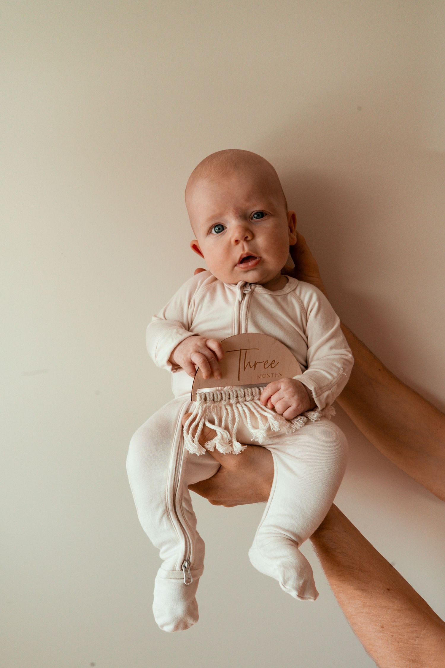 A baby in white sleepwear holds a brown card while lying on a neutral background.