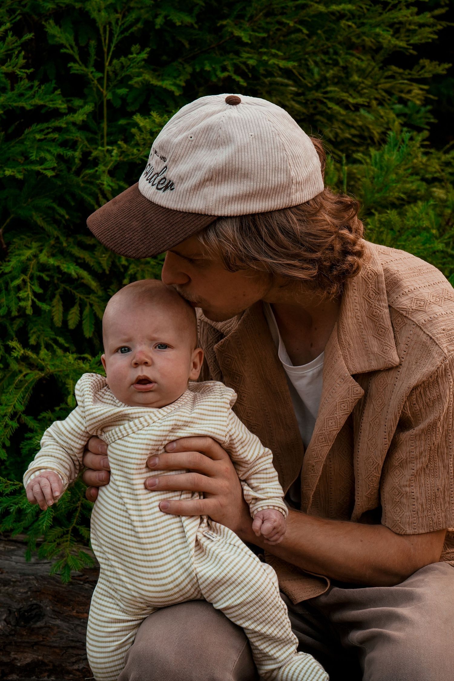 A caregiver holds and kisses a baby wearing striped clothing against a backdrop of evergreen trees.