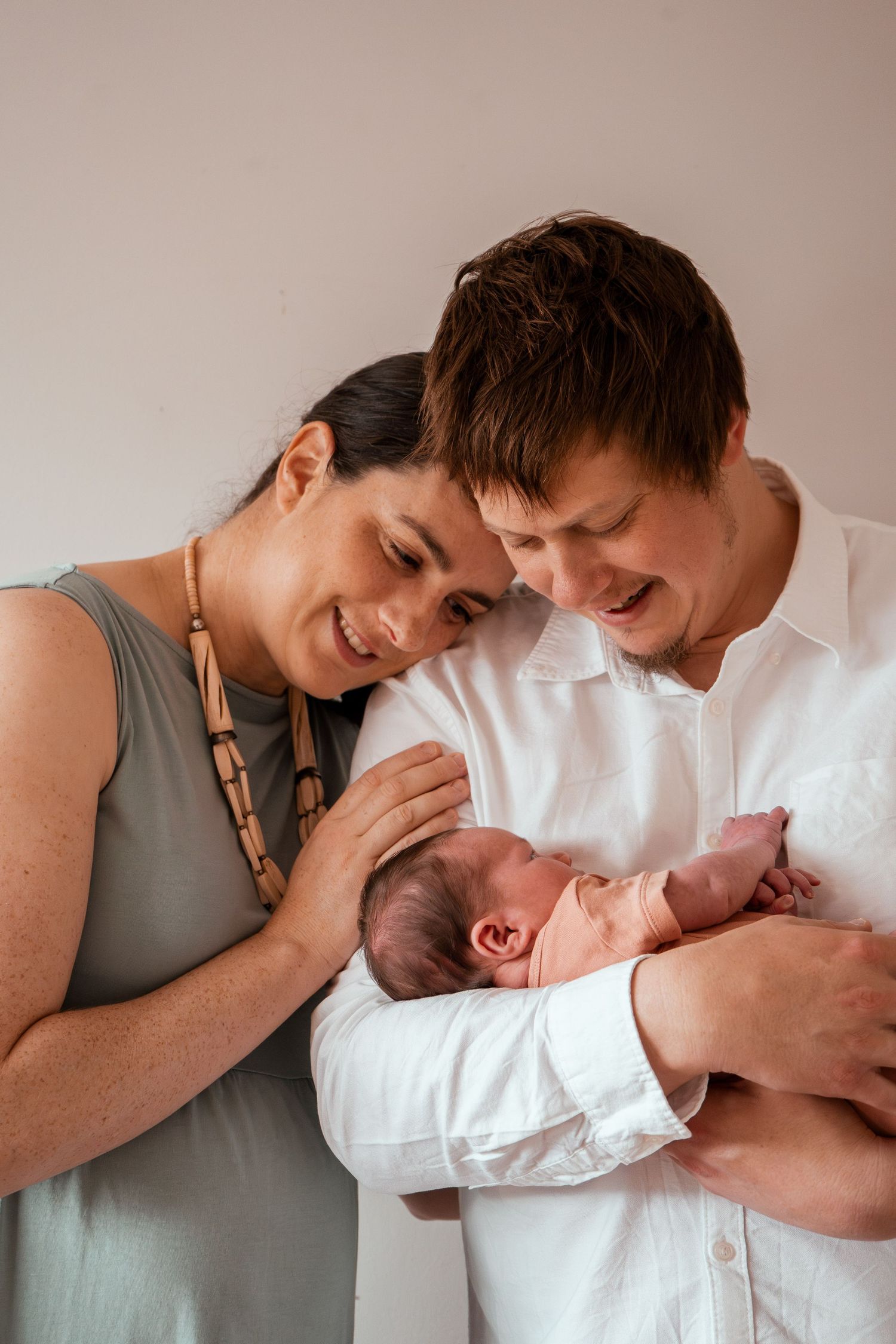 Parents in white and sage green attire lovingly gazing at their newborn baby during an intimate family moment.