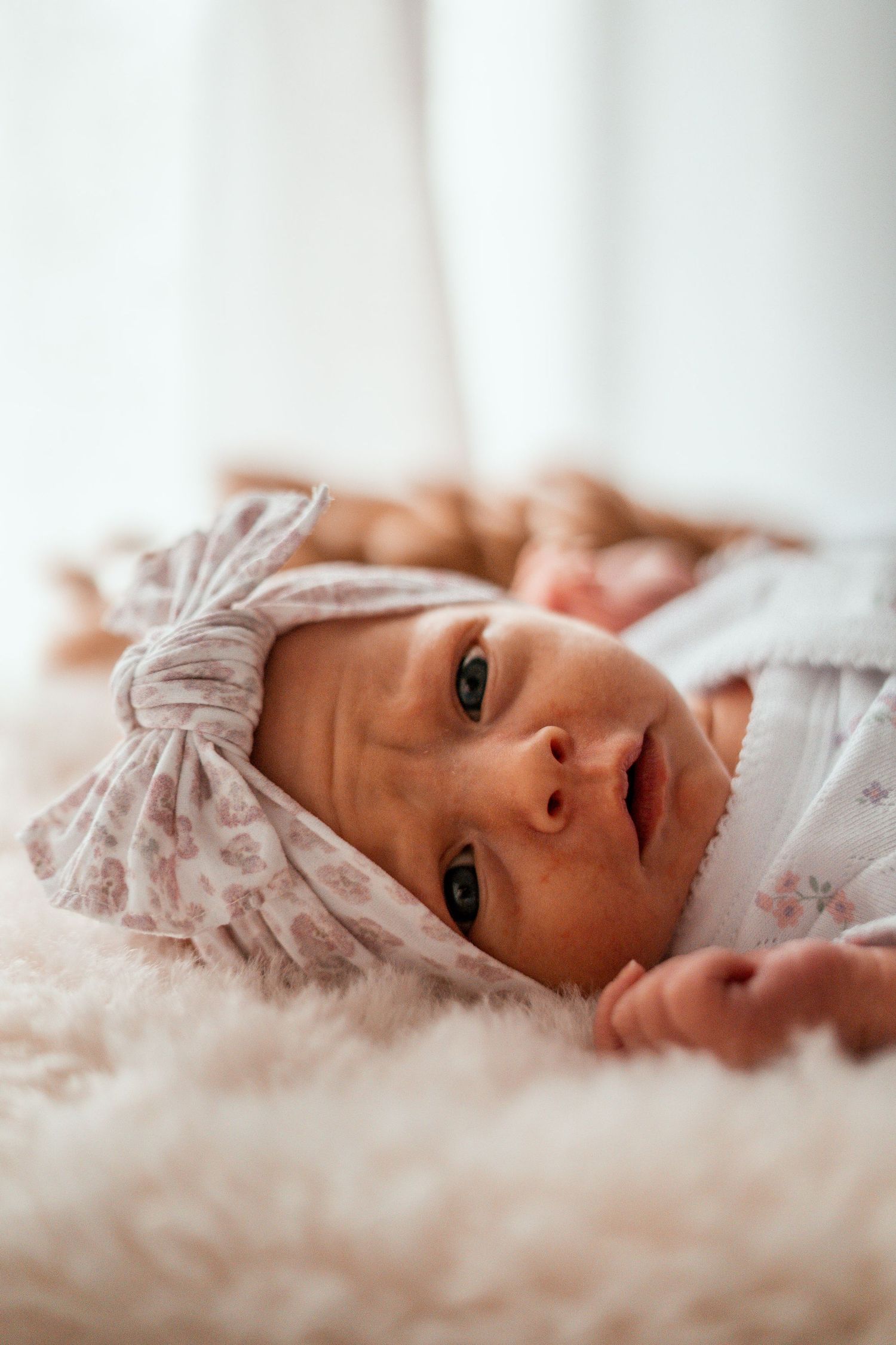 Baby wearing pink bow headband lying on soft white bedding in natural light.