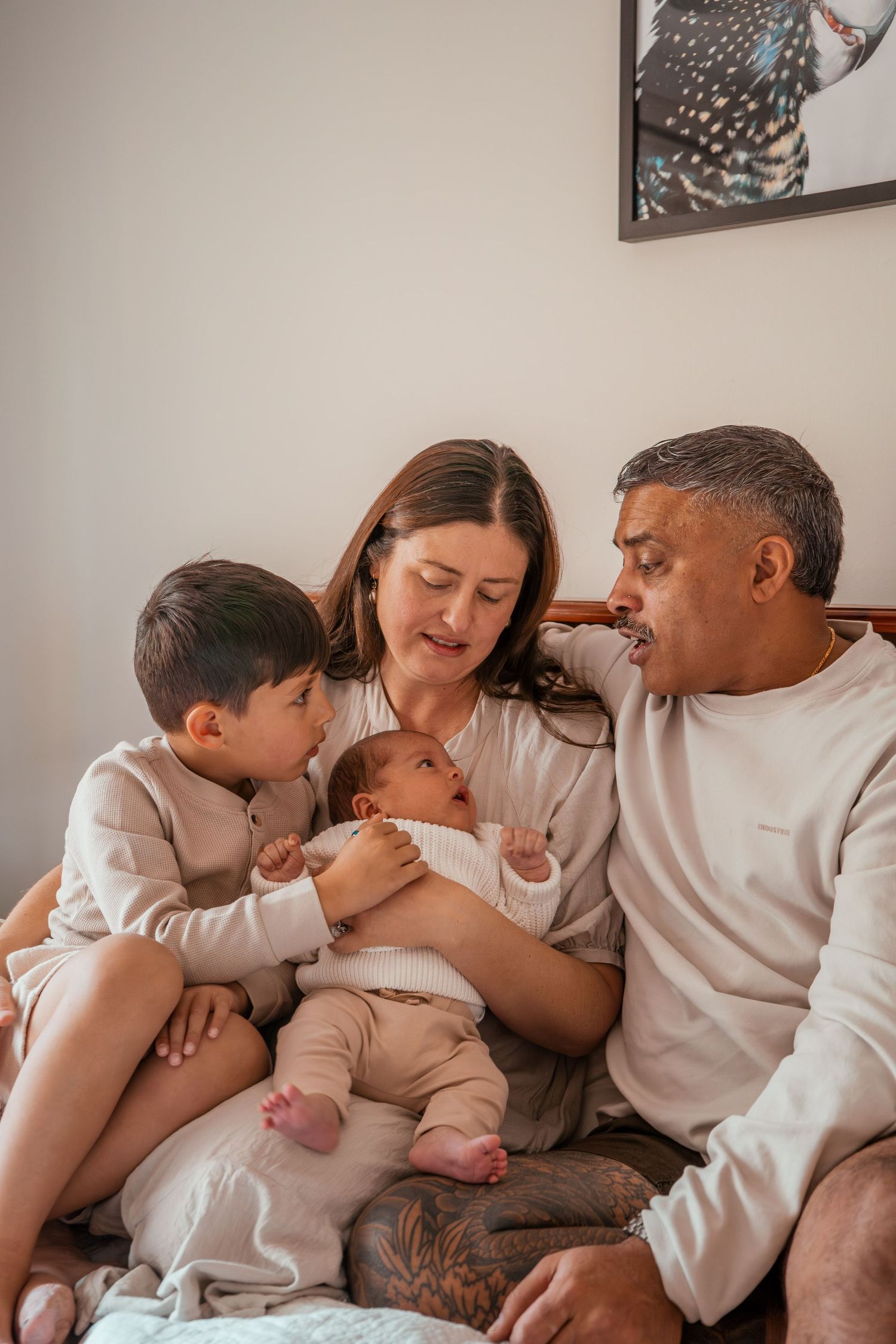 A family sits together on a bed in white clothing while admiring their newborn baby.
