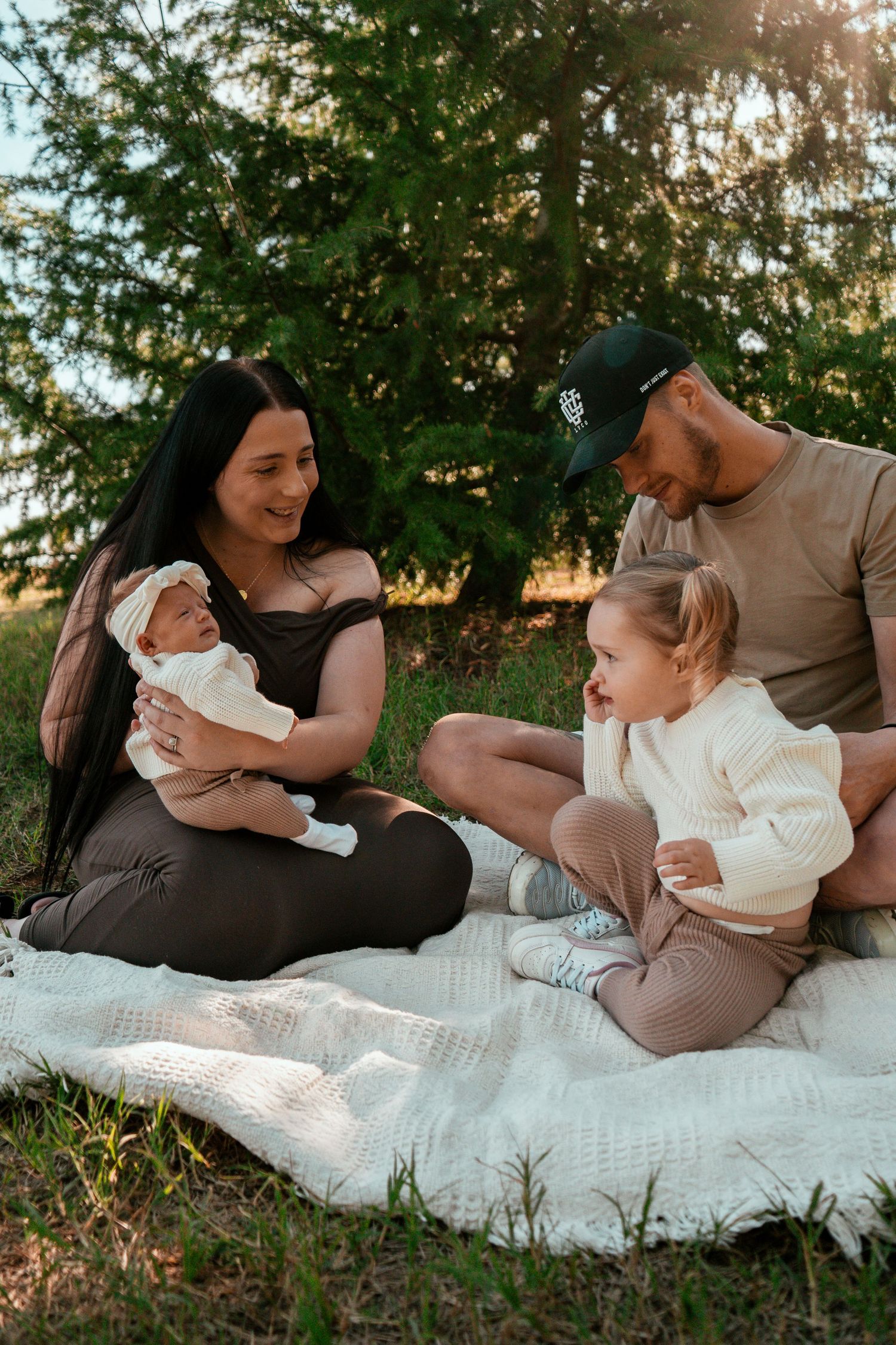 Family having a picnic outdoors on a white blanket with evergreen trees in the background during golden hour.