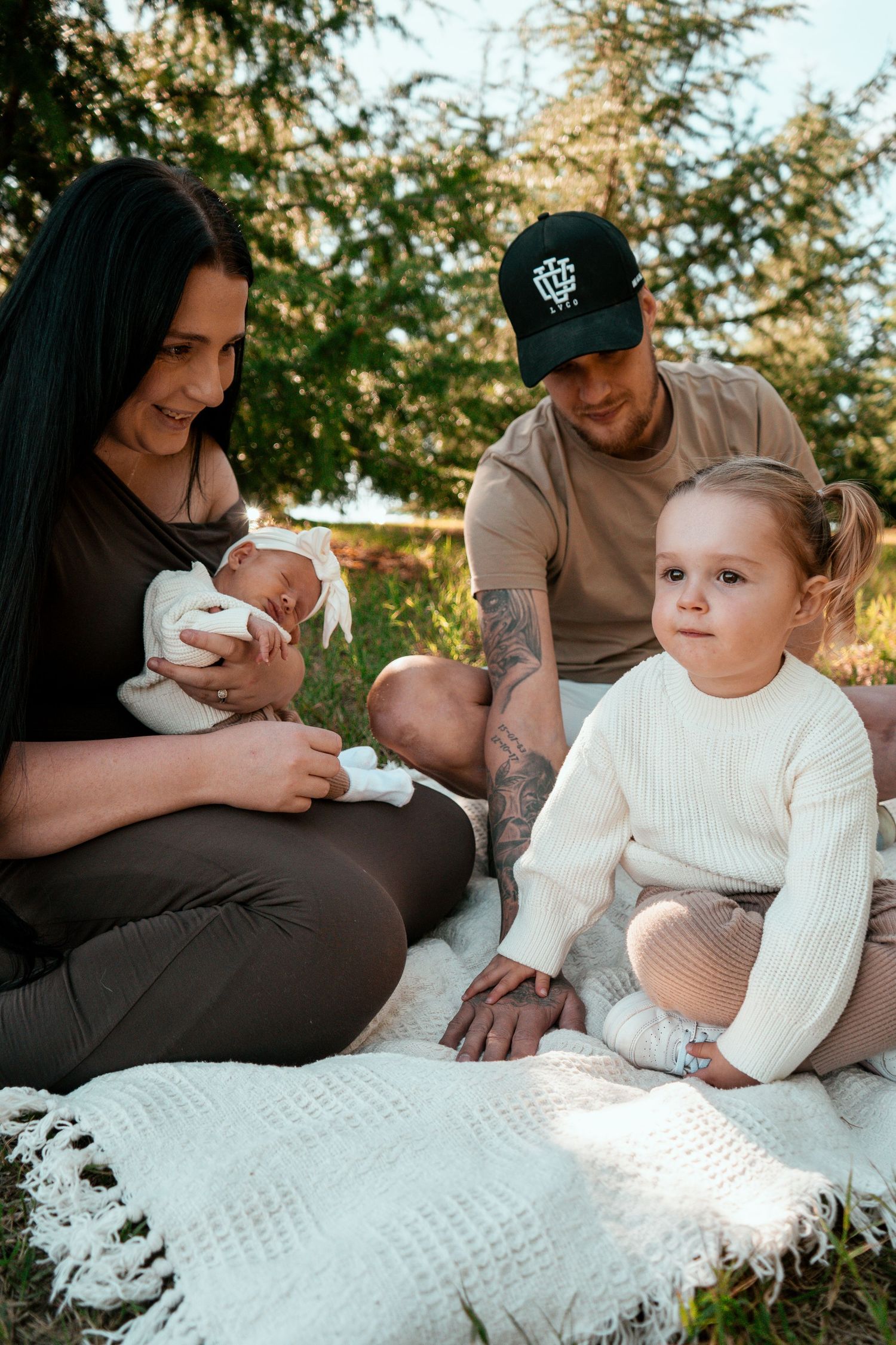 A family sitting together on a white blanket during an outdoor photo session in a wooded setting.