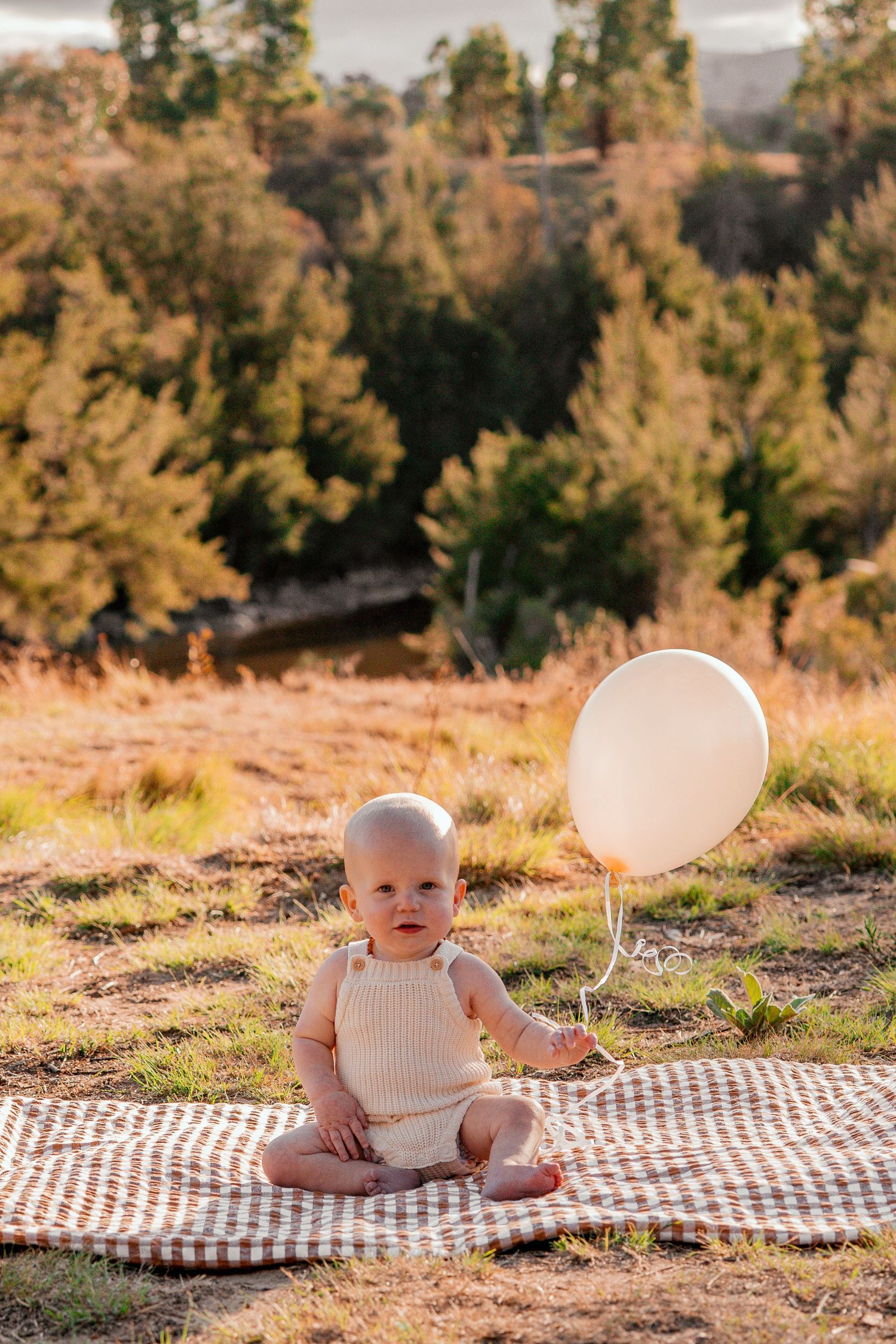 A baby sits on a checkered blanket outdoors during golden hour with white balloons in a natural setting.