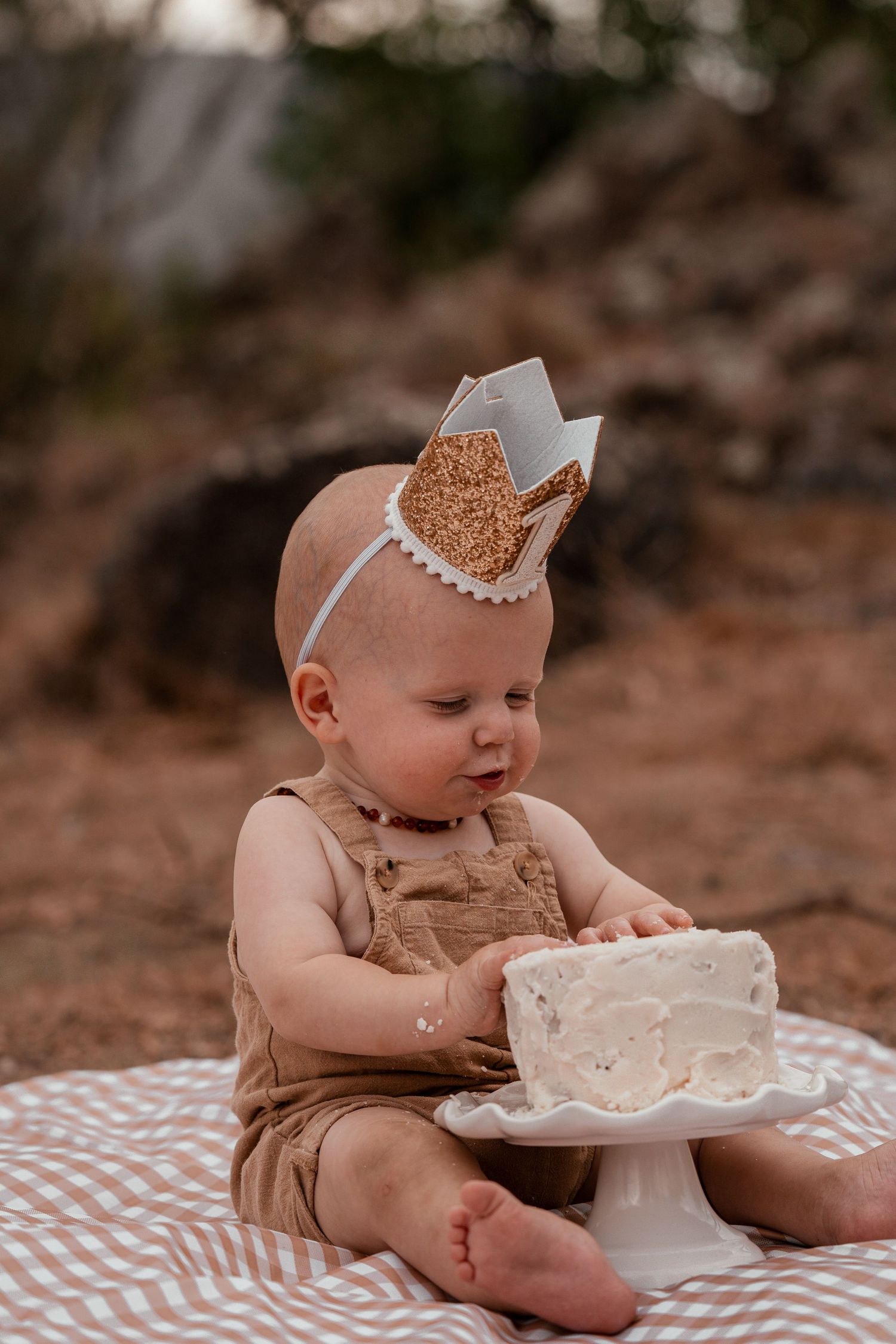 Baby wearing brown overalls and glitter crown sits on blanket with white cake for first birthday celebration.
