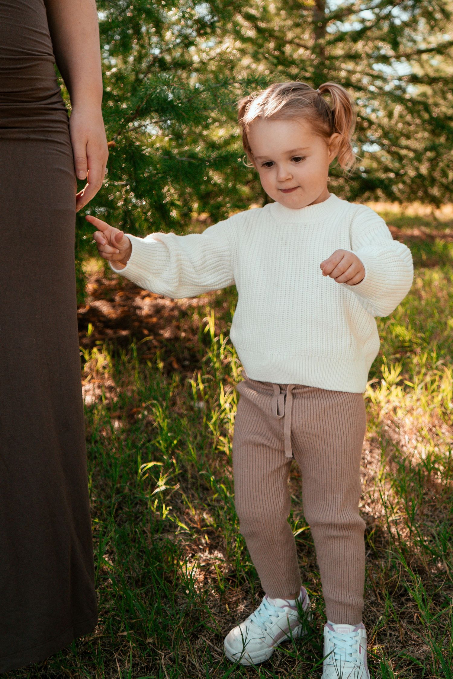 A small child in a white sweater and tan pants stands outdoors next to an adult.