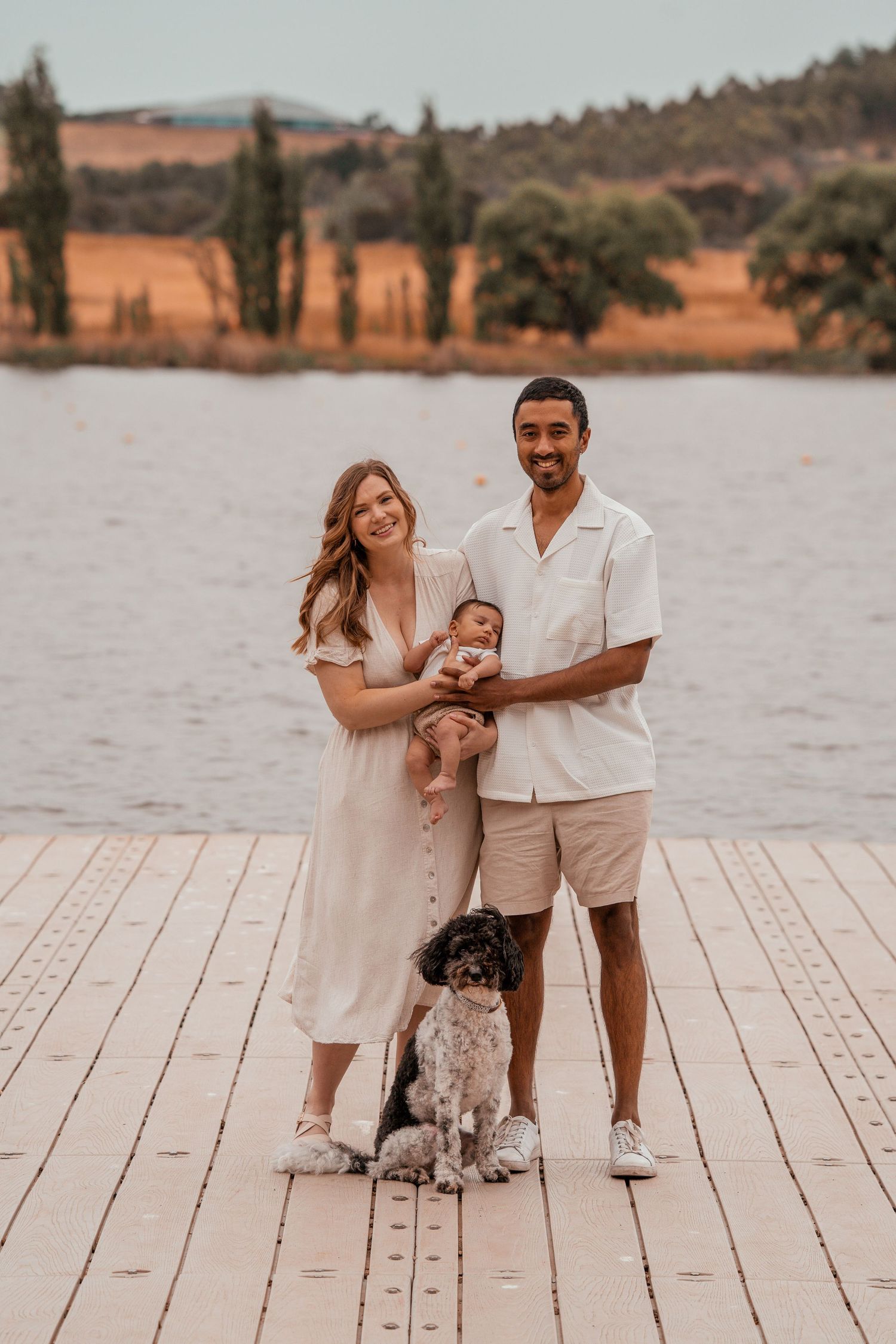 A family poses together on a wooden dock by a lake at sunset in coordinating neutral-colored outfits.
