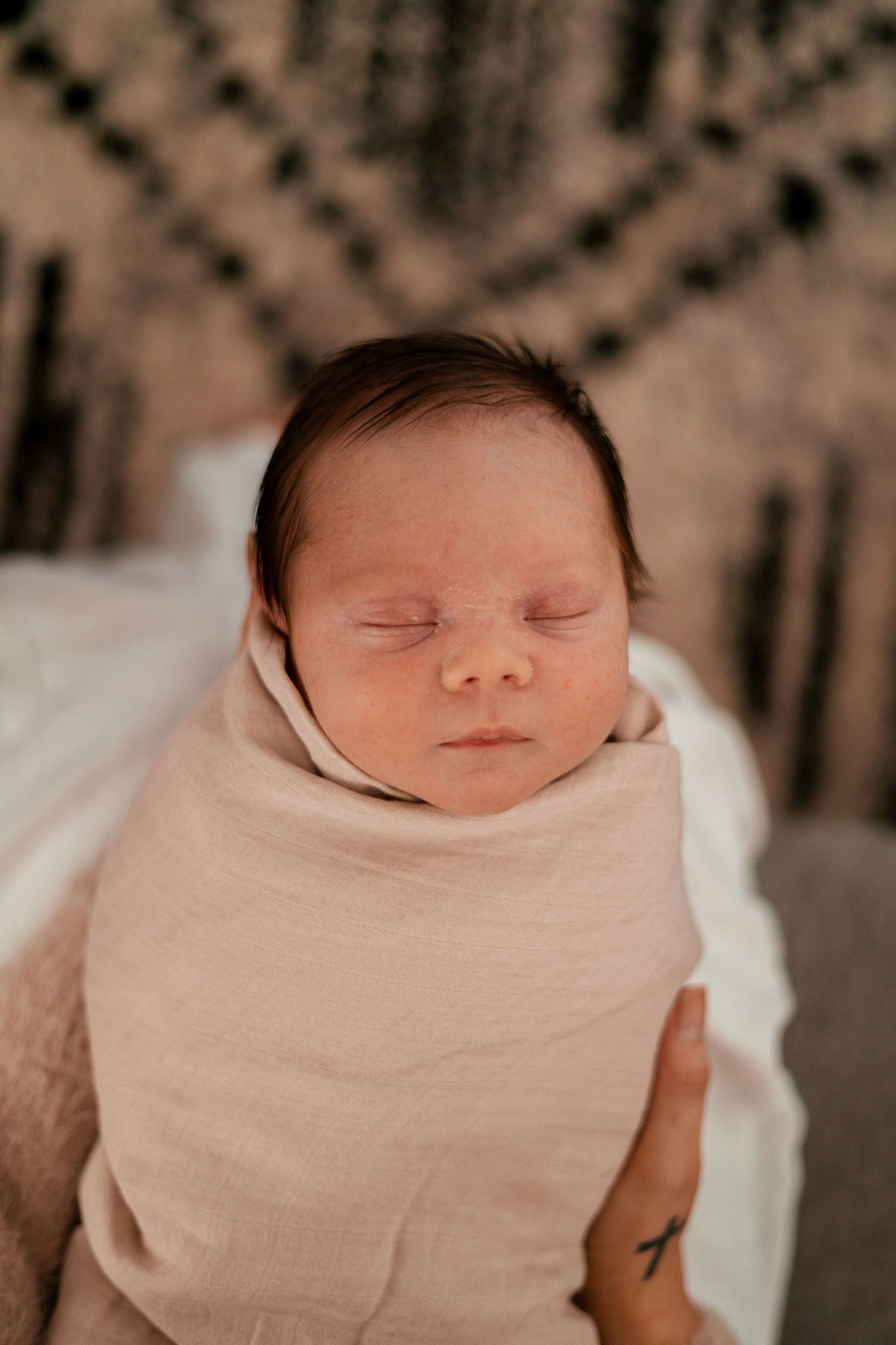 Sleeping newborn wrapped in a soft beige swaddle blanket against a patterned background.