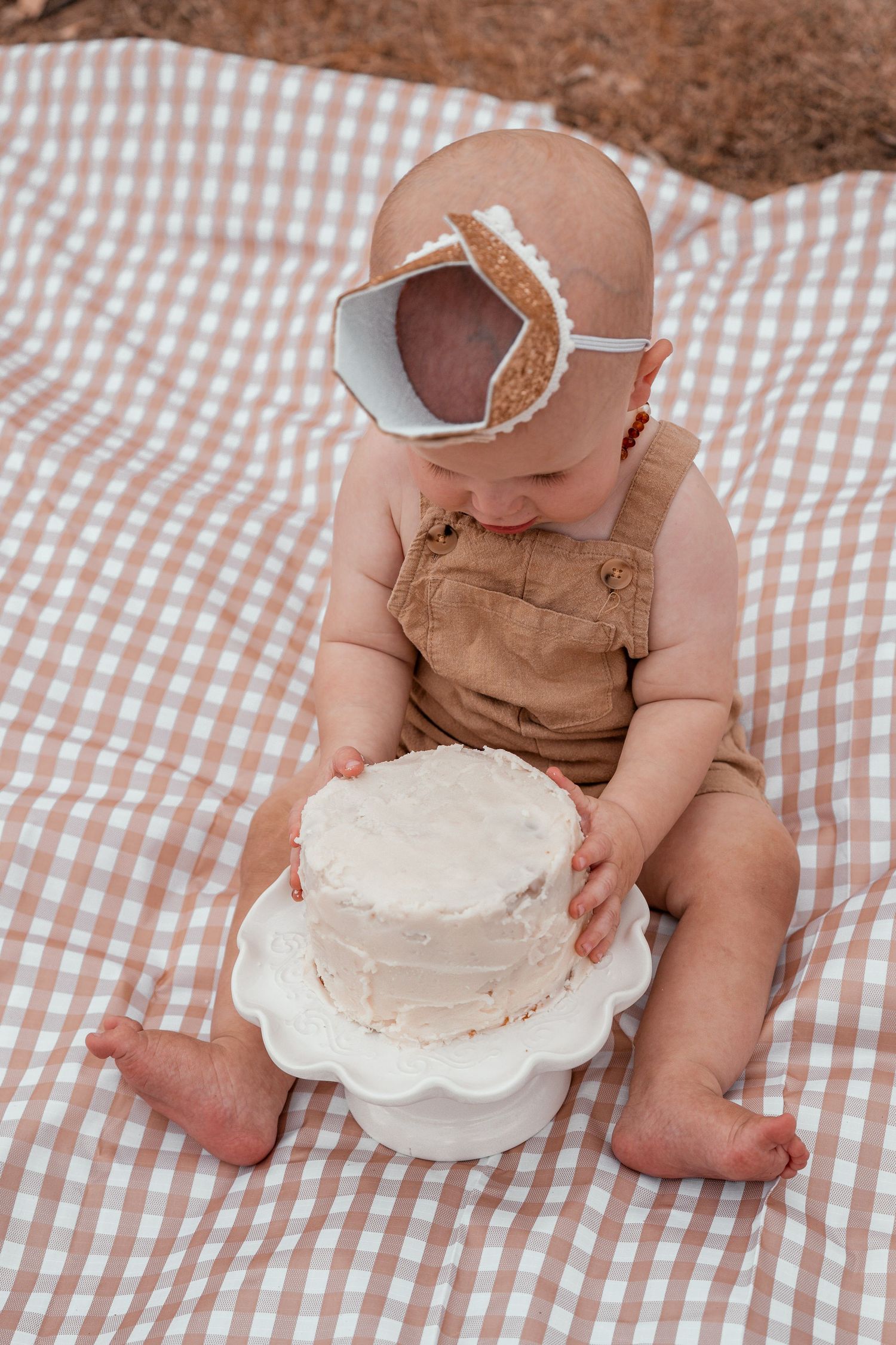 Baby in brown overalls explores white frosted cake during cake smash photo session on gingham blanket.