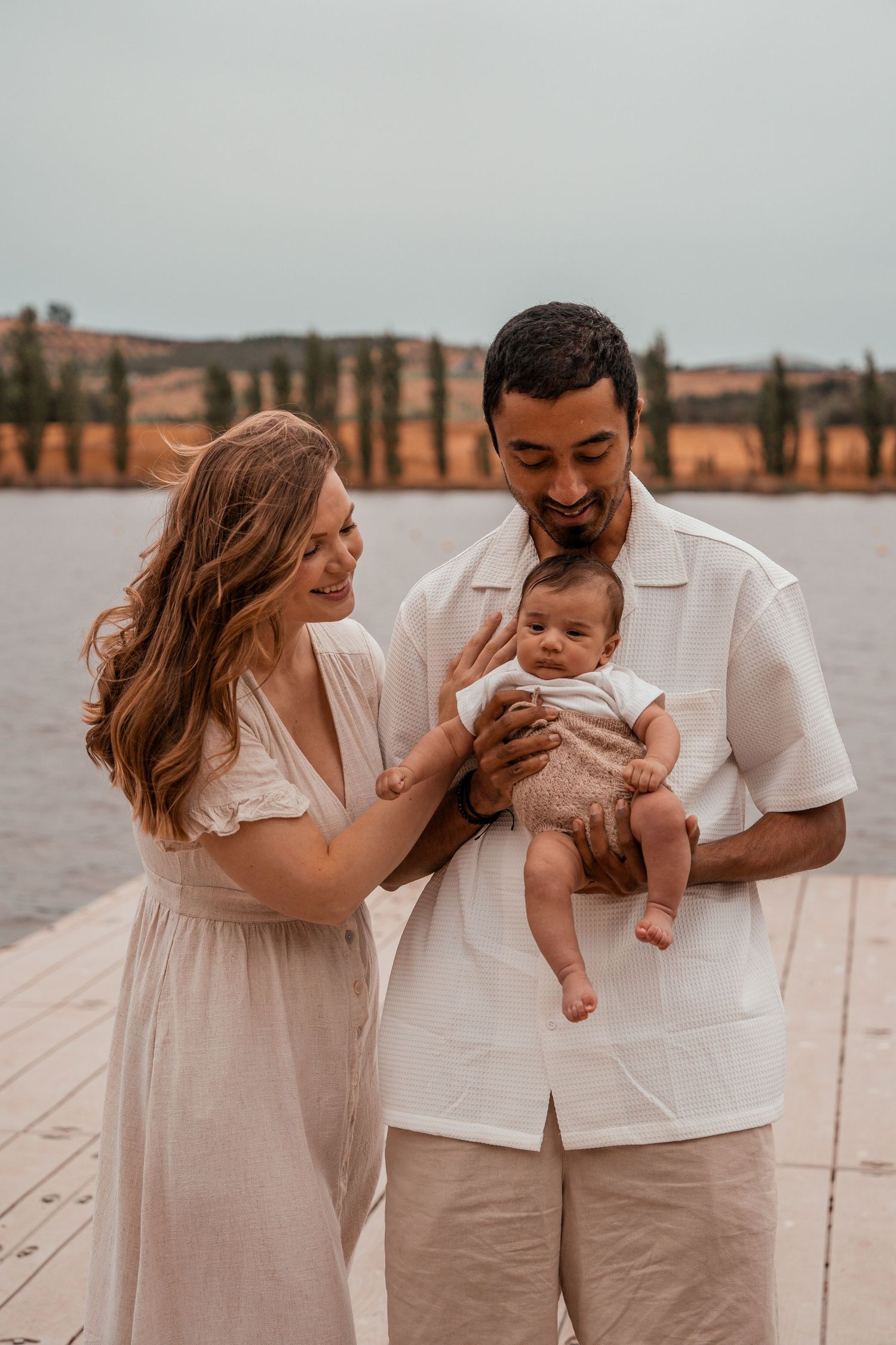A family in white and beige outfits shares tender moments with their baby on a wooden deck by the water.