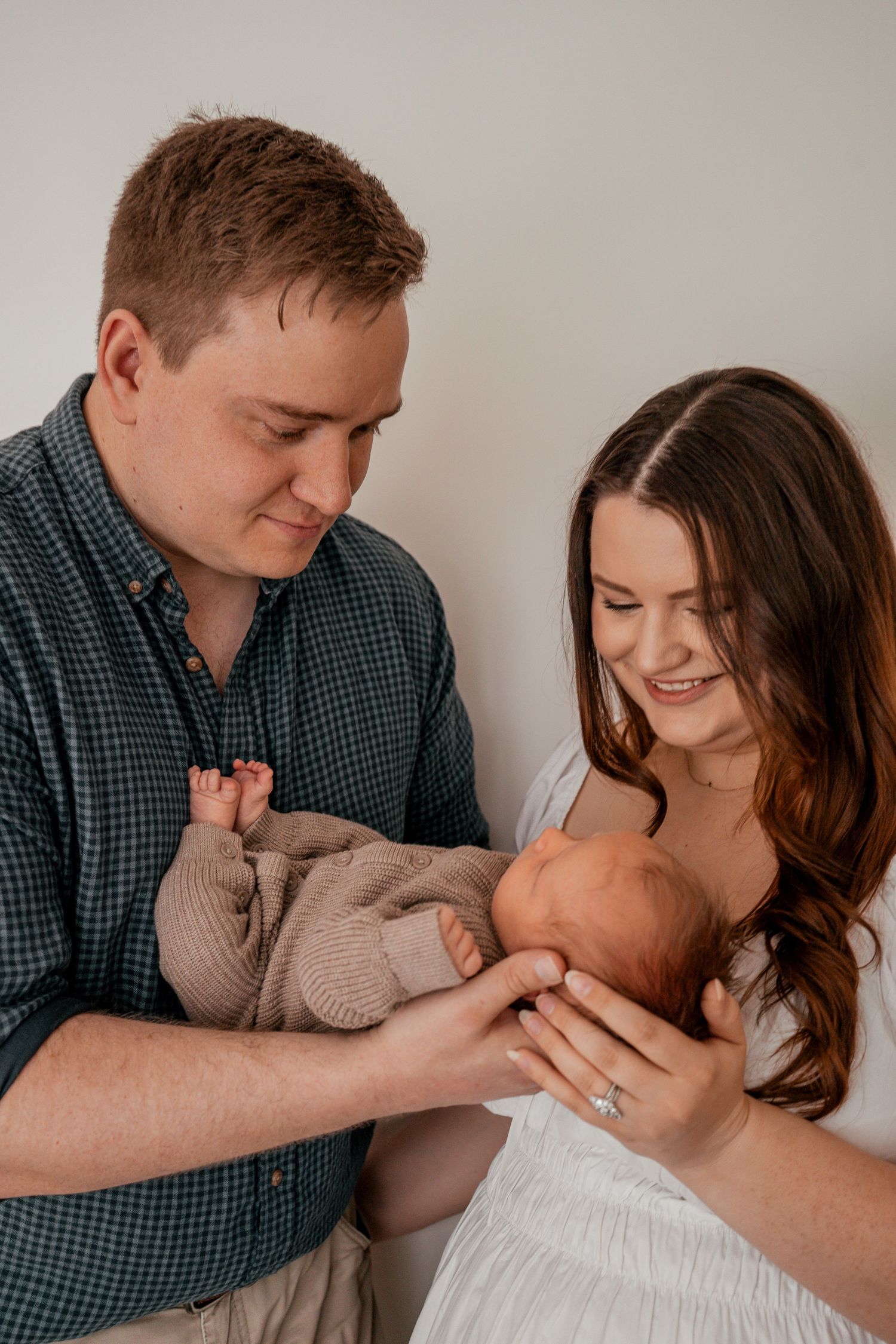 A couple lovingly gazes at their newborn baby wrapped in a beige sweater against a neutral background.