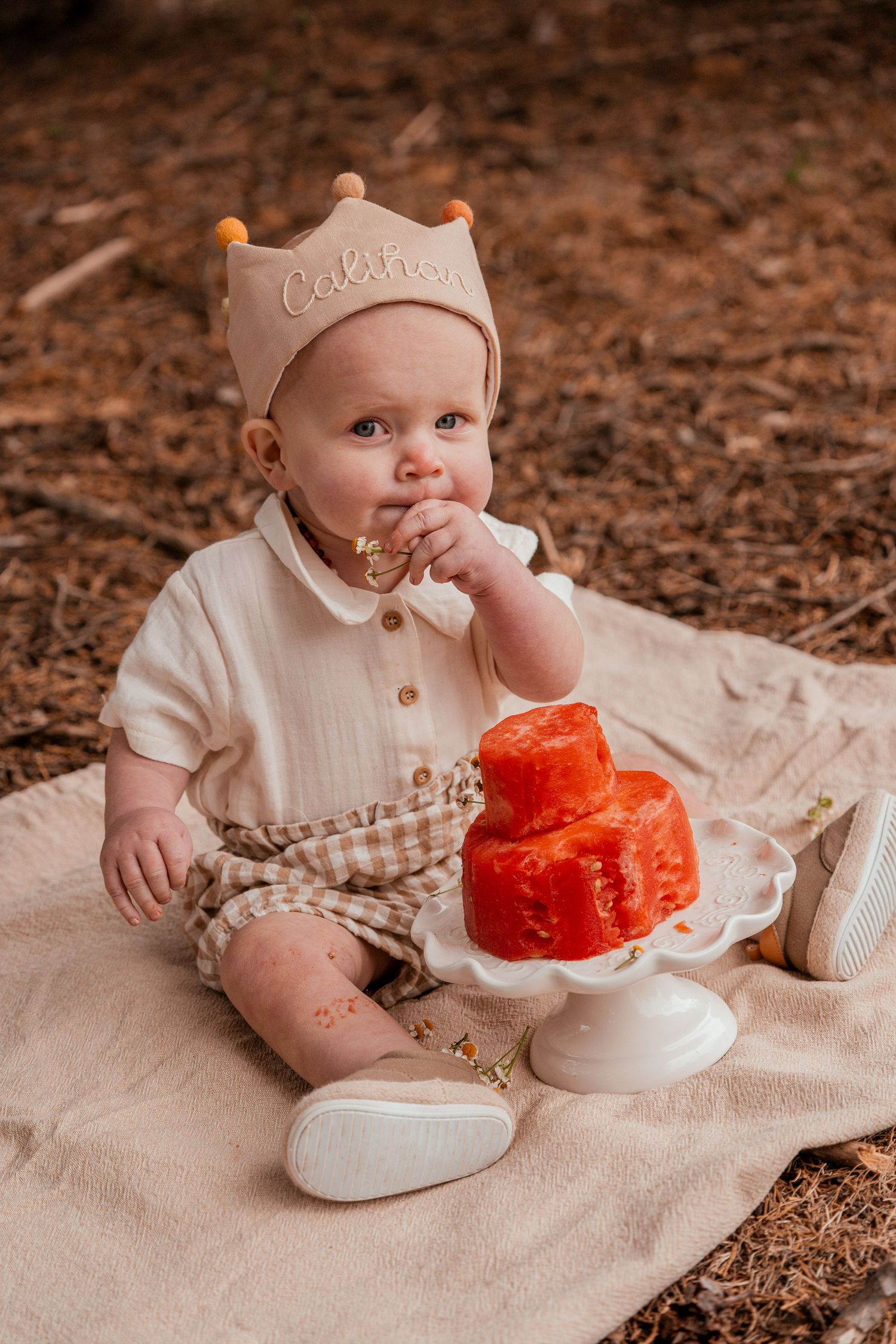 Baby in cream colored outfit sits on blanket with red smash cake during first birthday photo session.