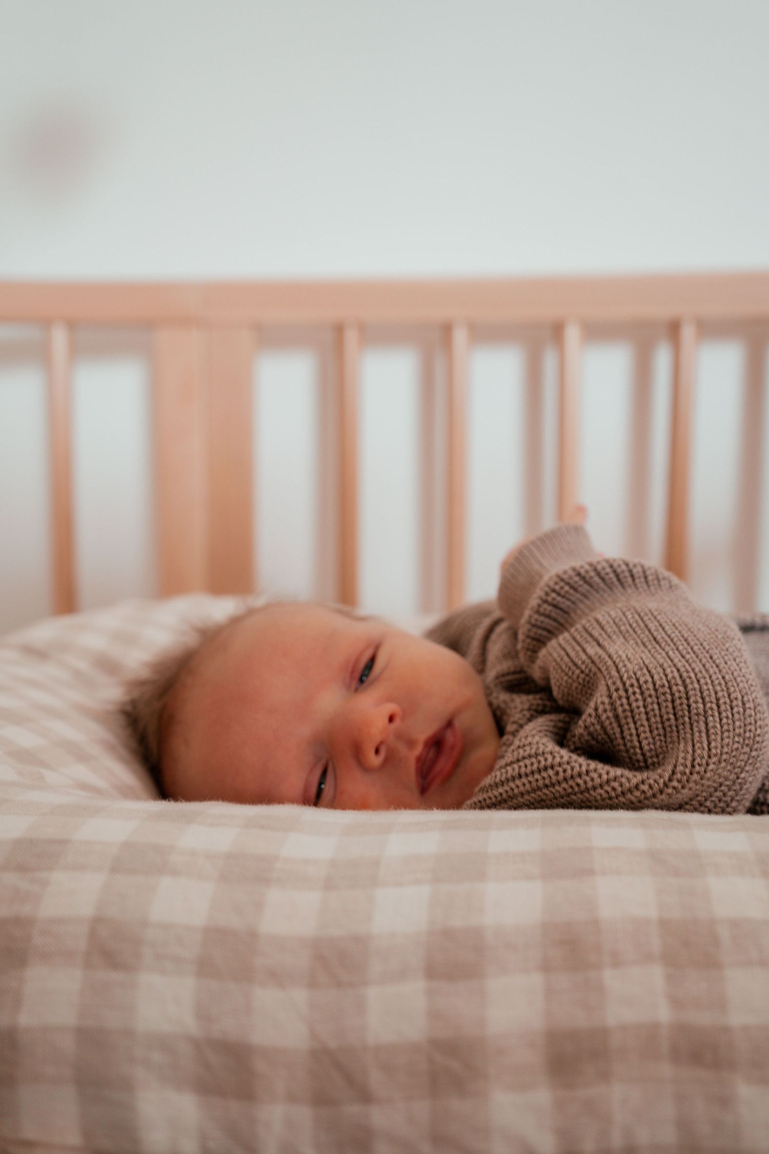 Newborn baby sleeping peacefully on plaid bedding while wearing a cozy brown knit sweater.