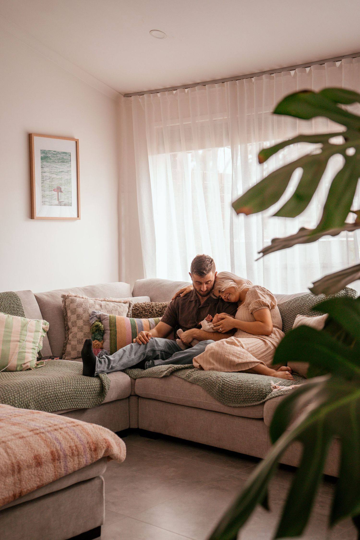 A relaxing scene with a couple and baby on sectional sofa near large windows and monstera plant.