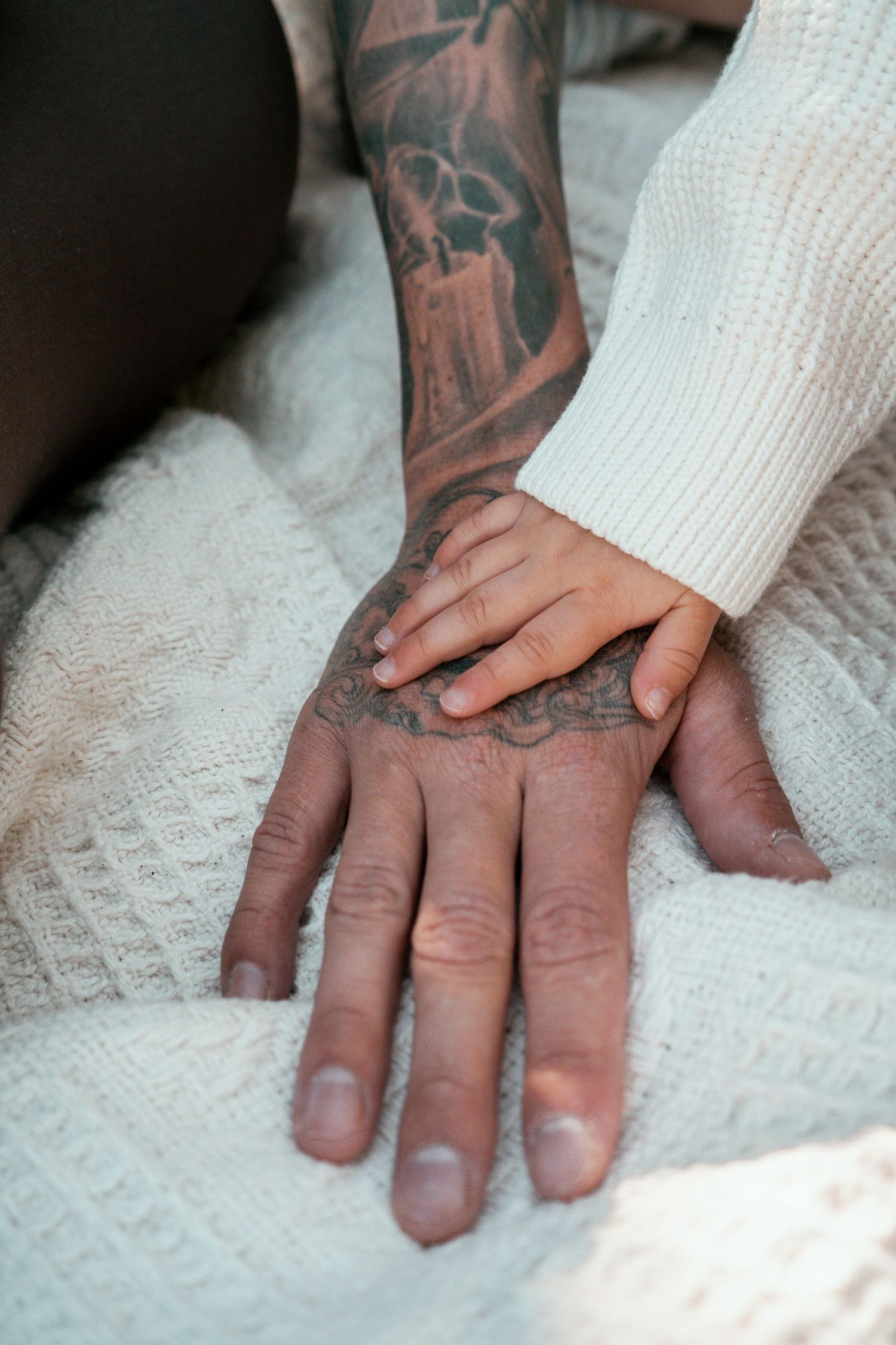 A small hand rests gently on top of a tattooed arm against a white textured background.