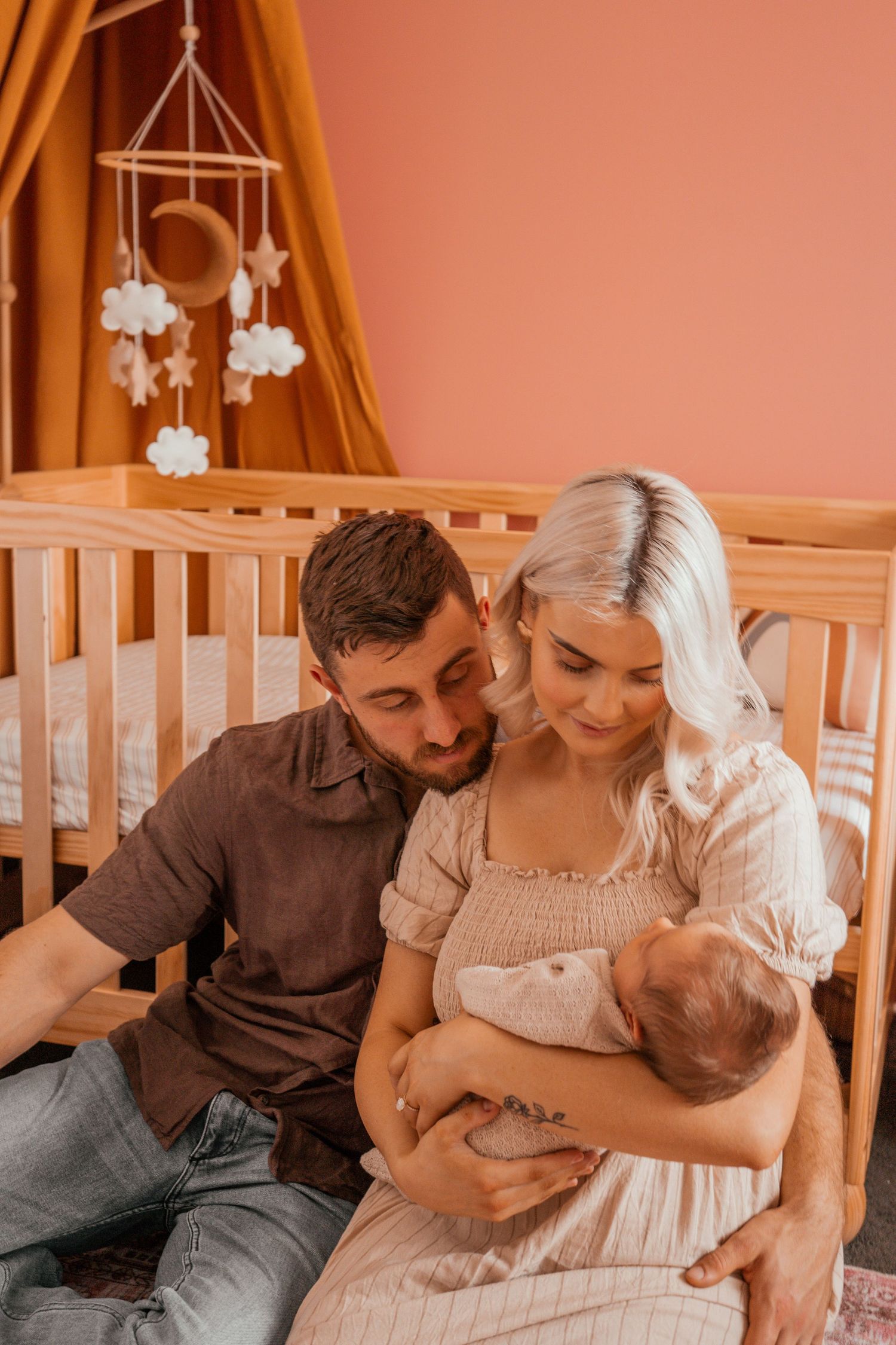 A family shares an intimate moment together in a nursery decorated with a cloud mobile and warm orange curtains.