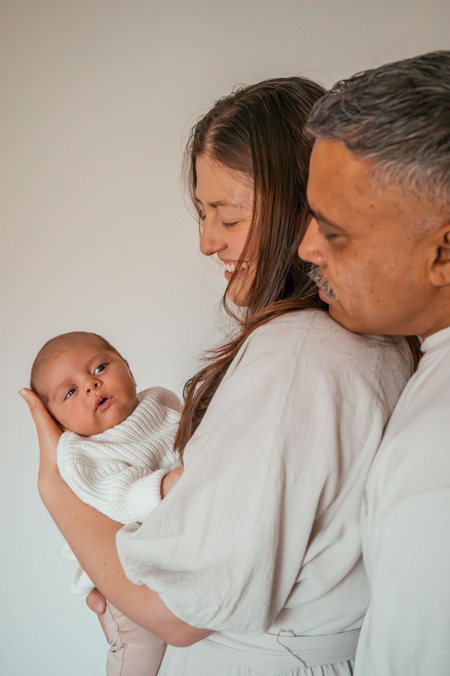 A couple in white robes tenderly holds their newborn baby in a warm, intimate family portrait.