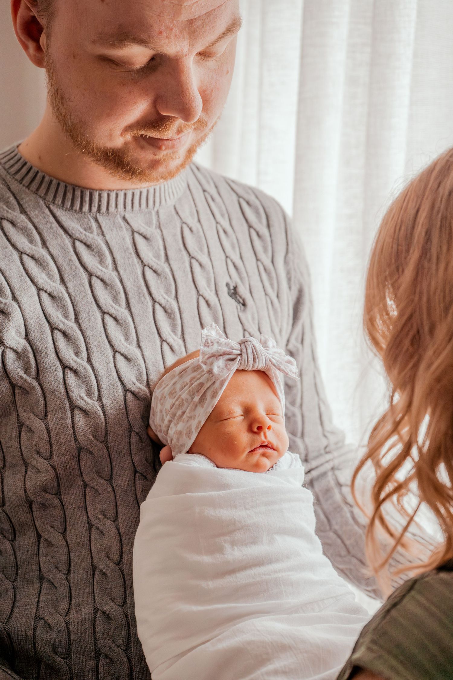 A newborn baby wrapped in a white blanket is held against someone wearing a gray cable knit sweater.