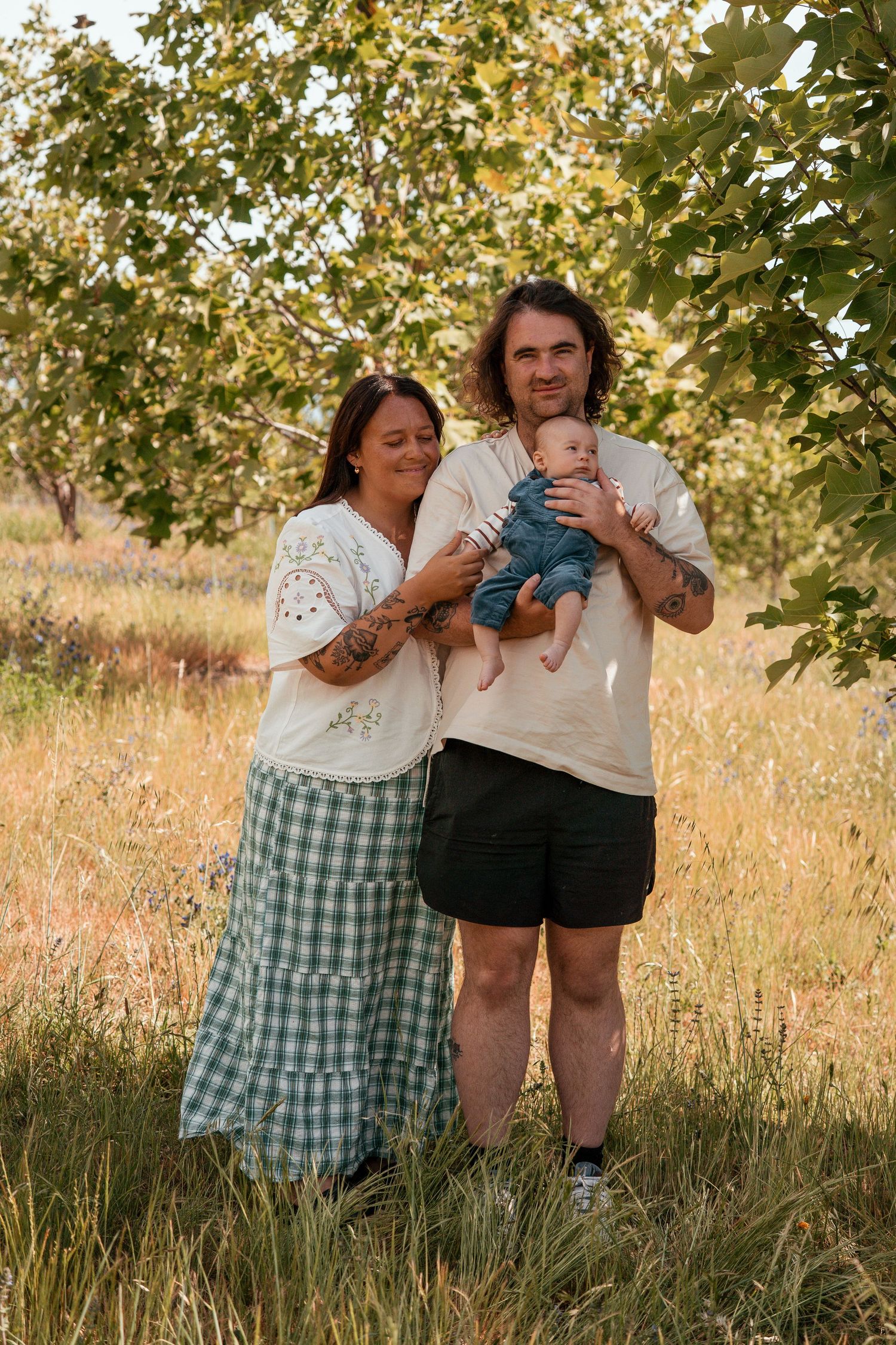 A family shares tender moments together in a sunlit field with tall grass and trees in the background.