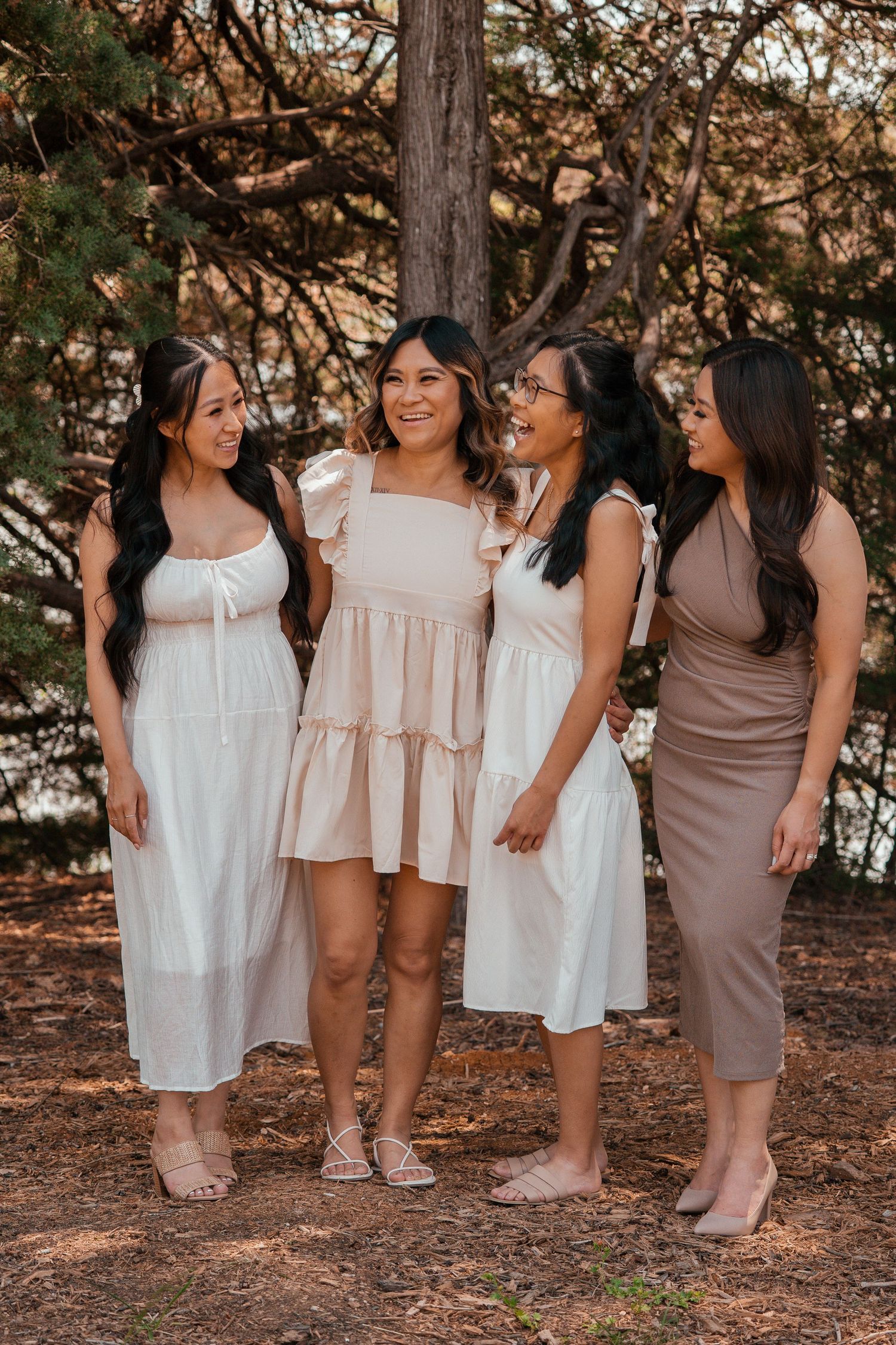Group of friends in neutral-toned dresses standing together on pine needles in a forest setting at sunset.