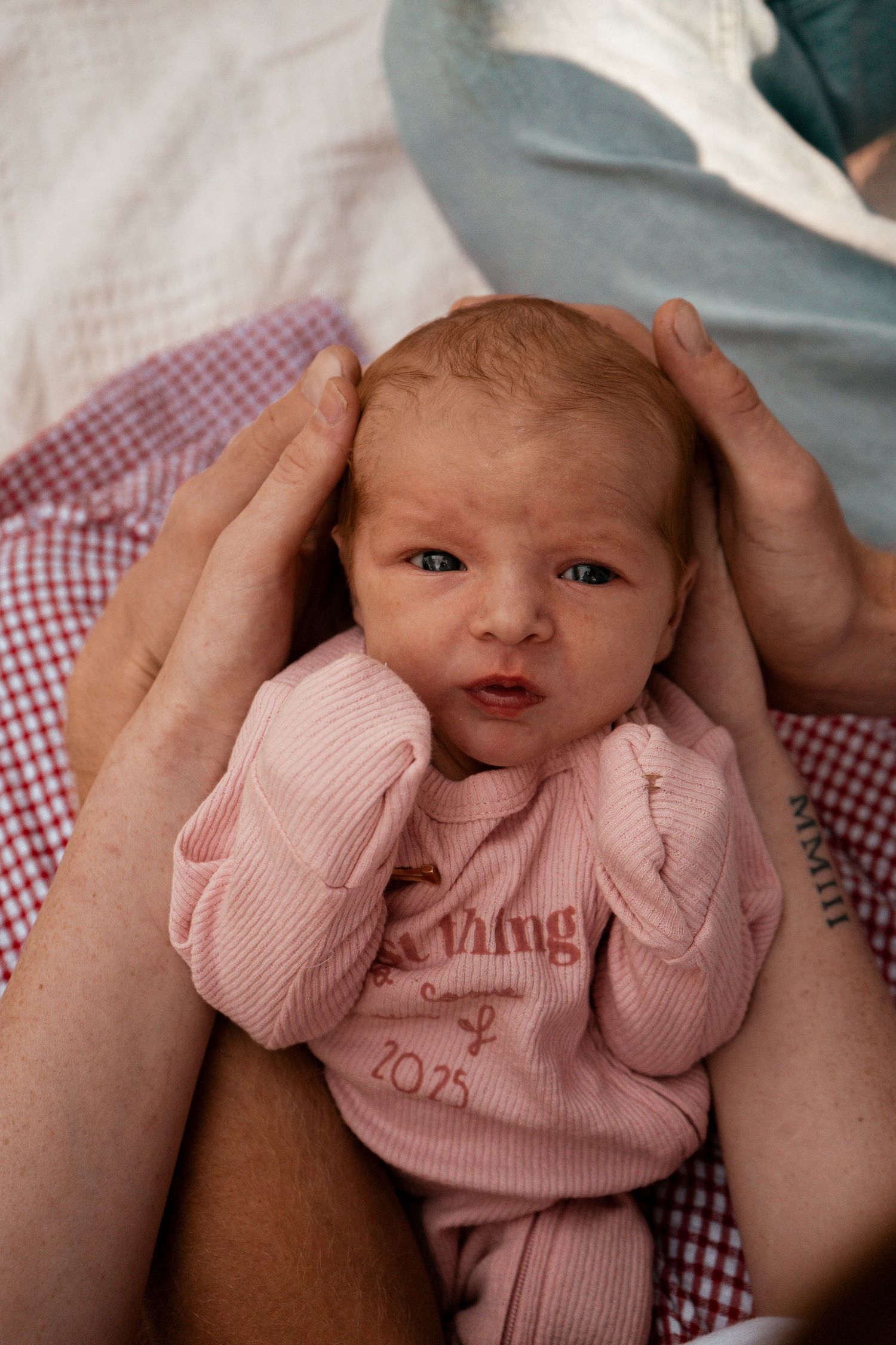 A small baby in pink clothing lies on a checkered blanket while being cradled.
