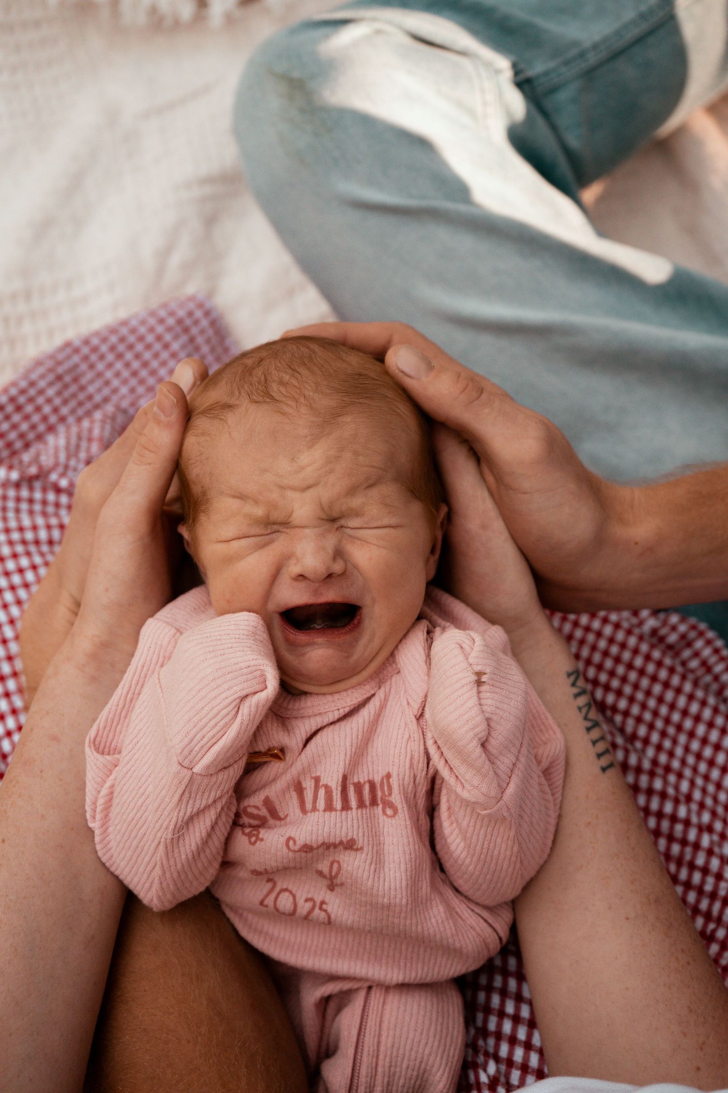 Baby in pink outfit making expressive crying face while being held.