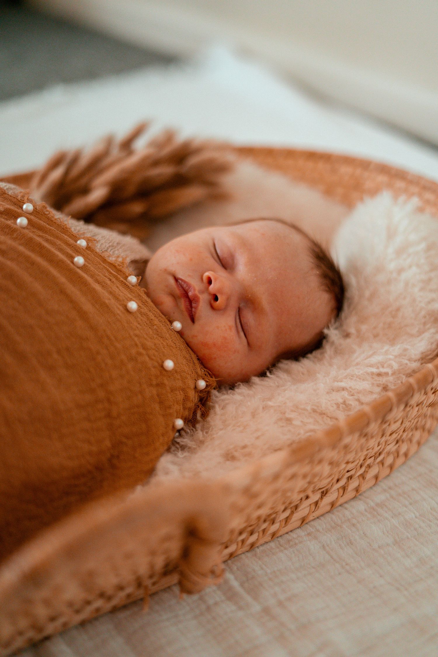 A sleeping baby lies peacefully on a plush blanket wearing a brown button-up outfit in soft natural lighting.