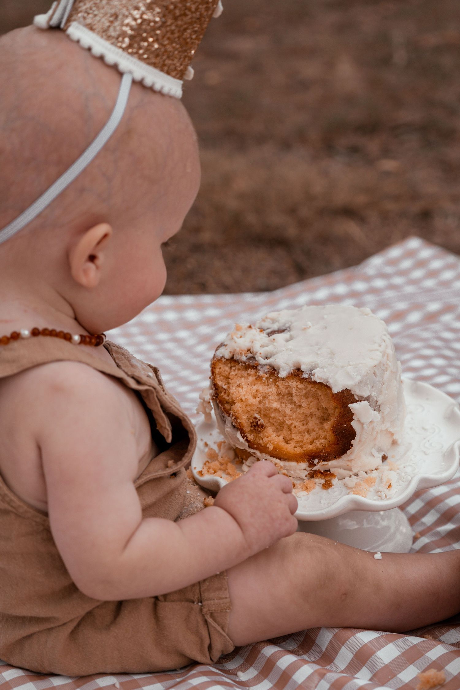 Baby in brown outfit enjoying a messy cake smash photo session on checkered blanket with frosted dessert.