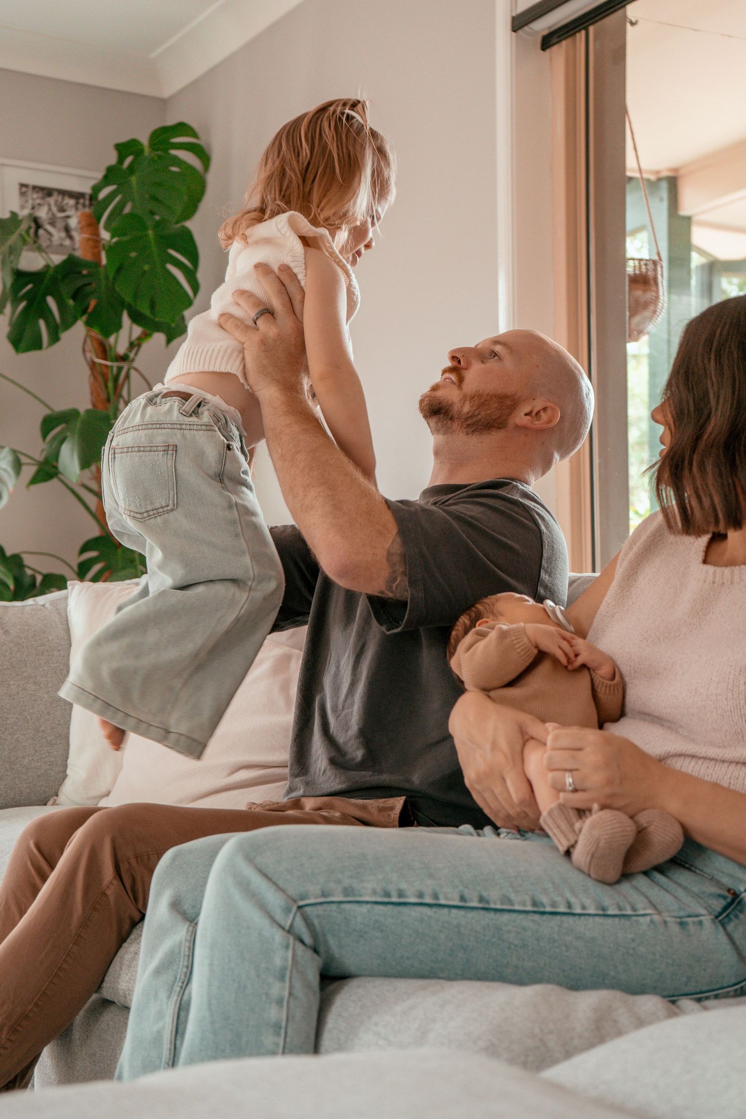 Family relaxing together on a couch in a bright living room with monstera plants in the background.