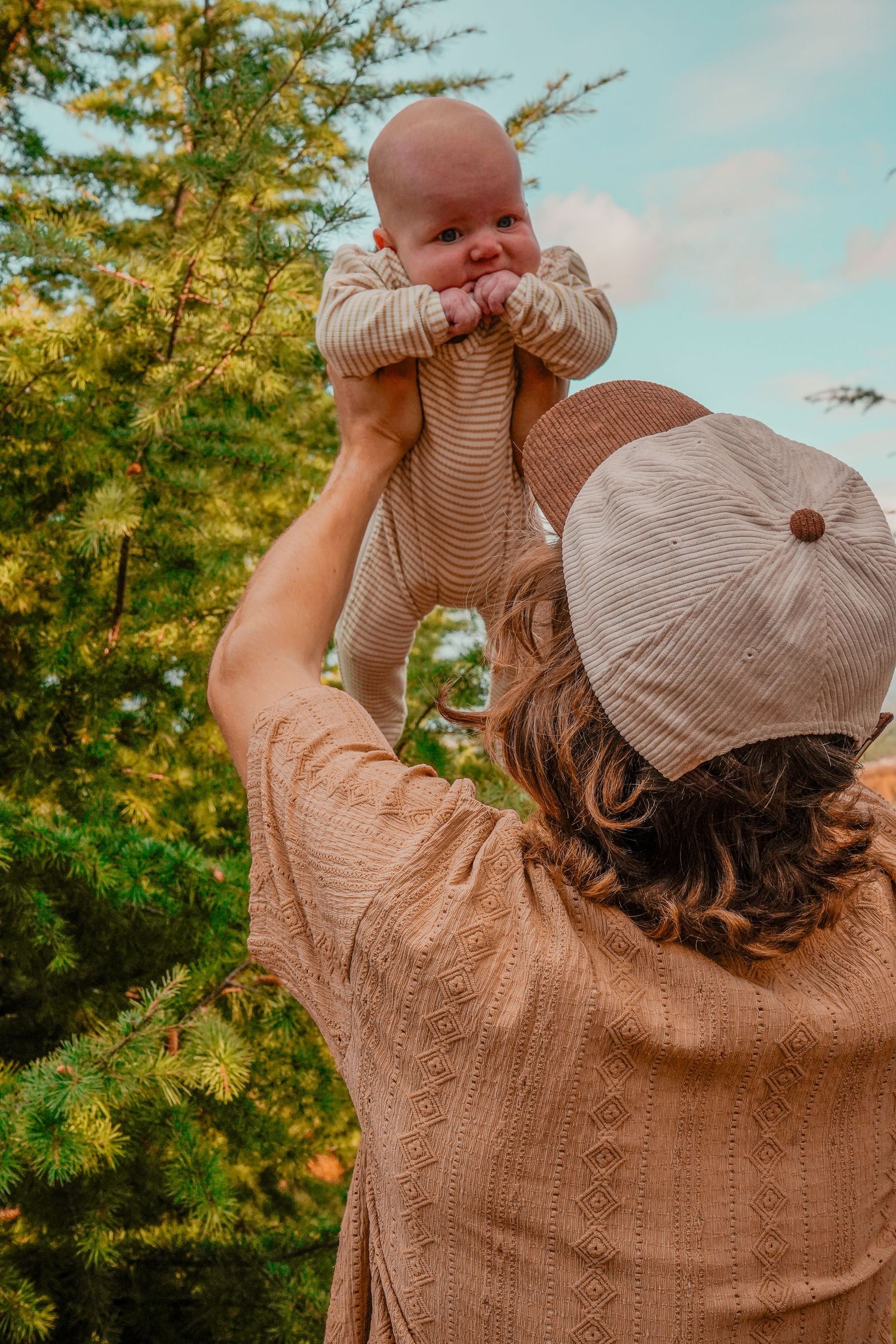 Someone in a brown sweater lifts a smiling baby against a blue sky and pine trees.