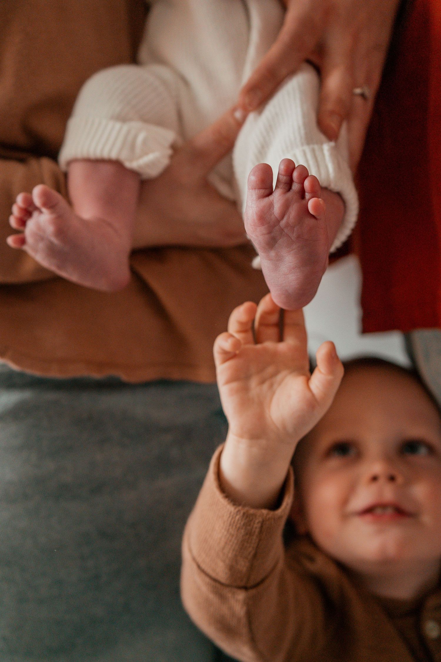 A toddler in brown clothing reaches up to touch tiny baby feet wearing white knit booties.