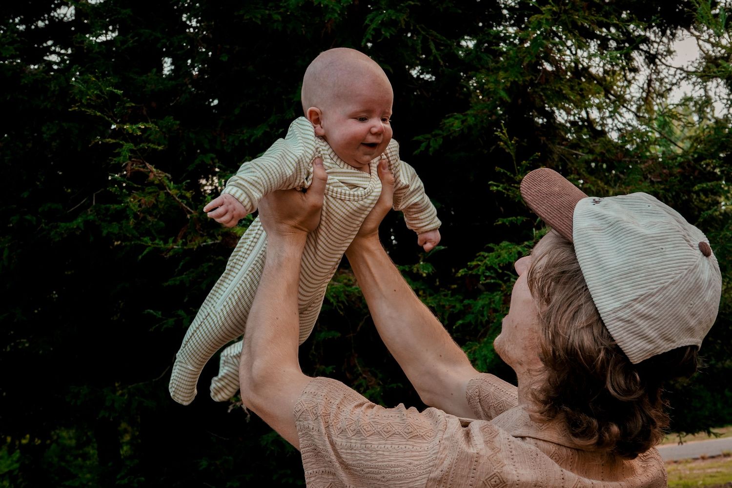 Parent lifting joyful baby in white onesie high in the air outdoors with trees in background.