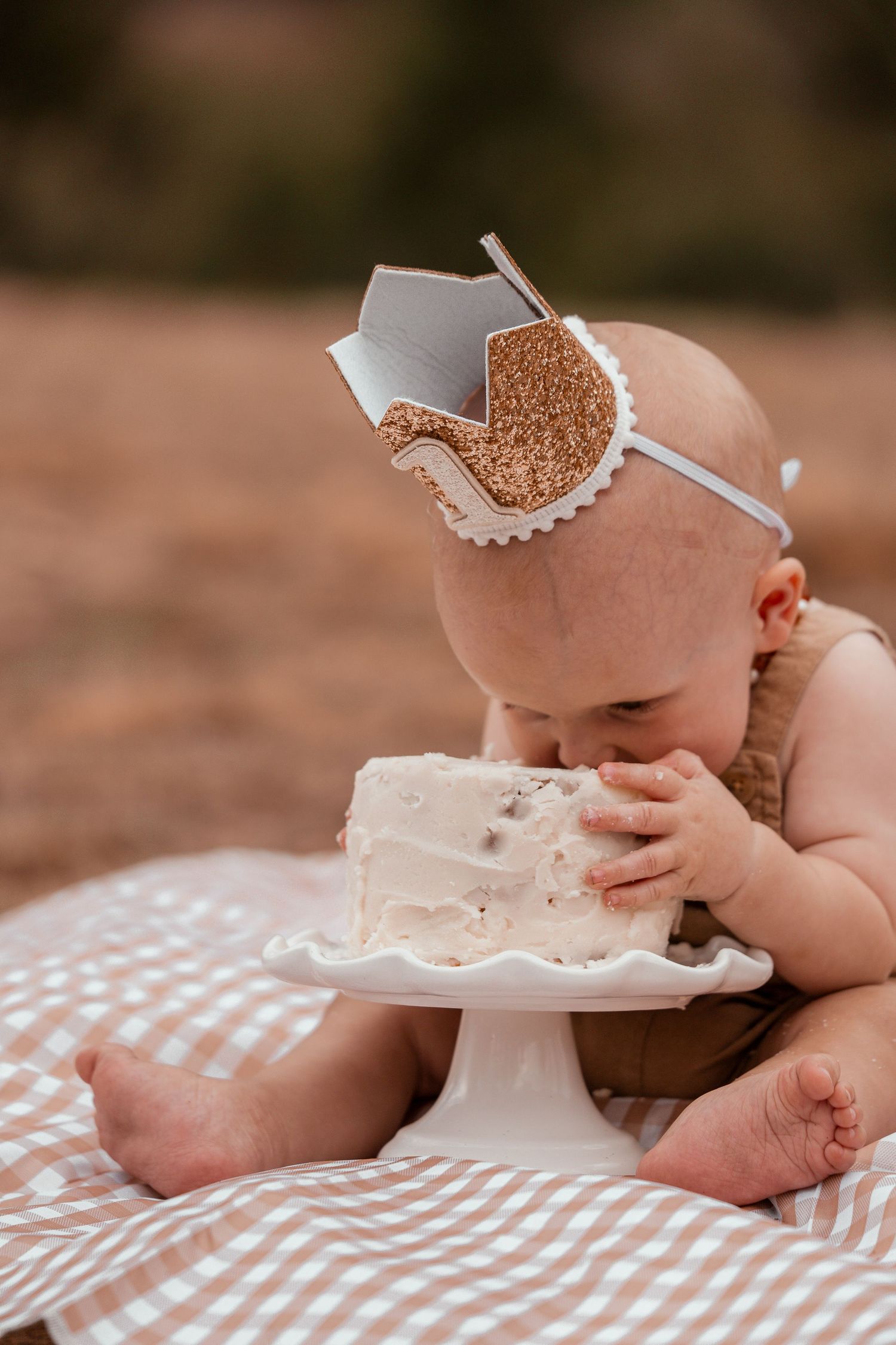 A baby wearing a white crown reaching for and eating a slice of birthday cake on a checkered blanket.