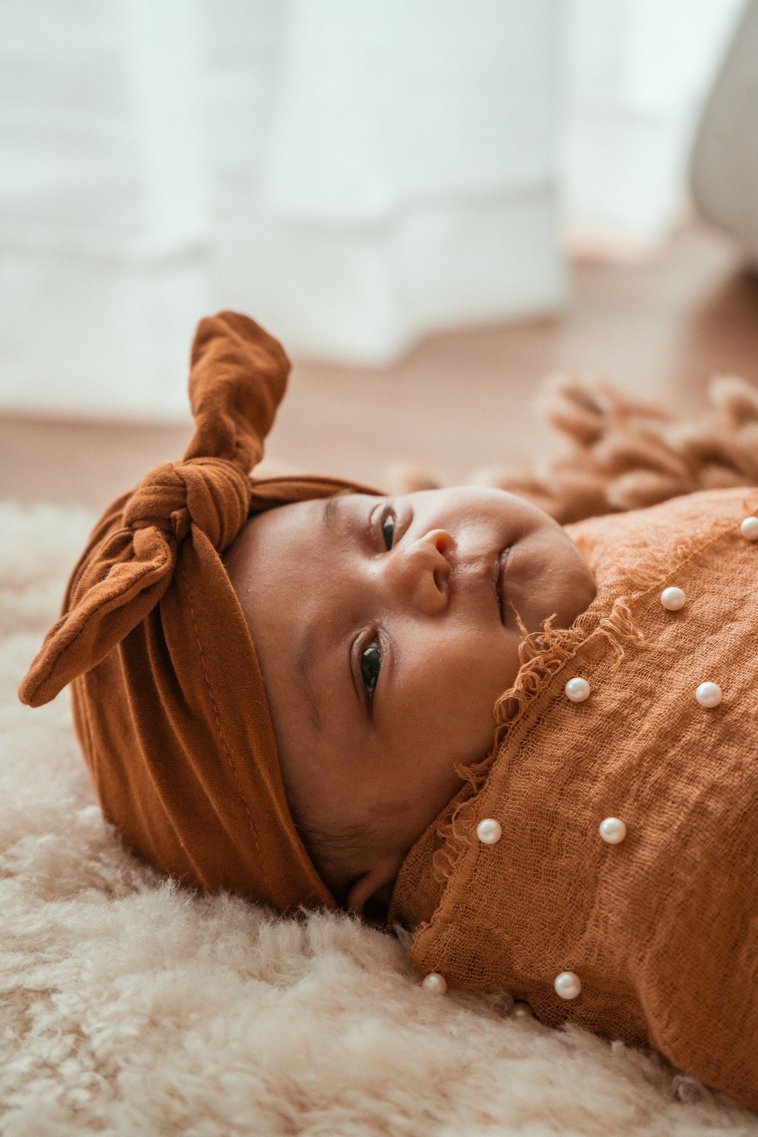 A baby wearing a brown headband and button-up outfit lies on a soft white fur blanket.