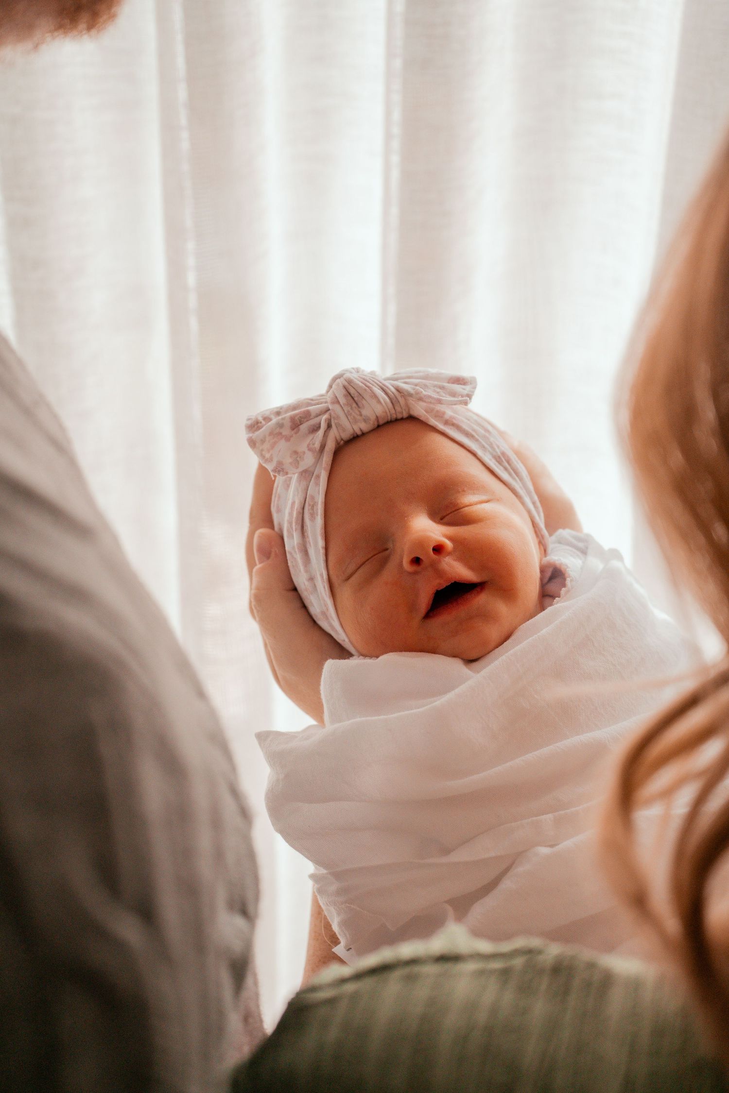 Peaceful sleeping newborn baby swaddled in white blanket against soft window light.