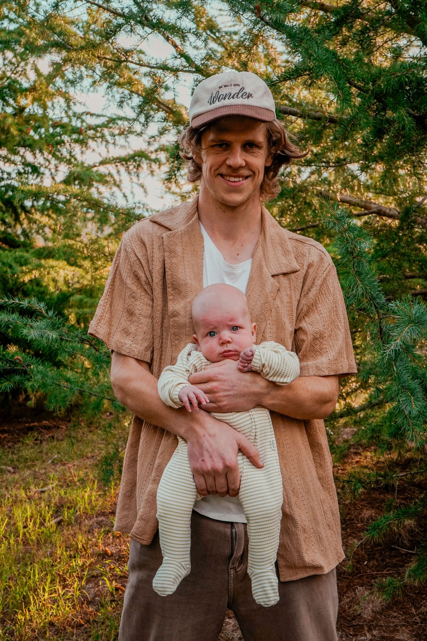 A tender outdoor family portrait showing a parent holding a small infant against a backdrop of sunlit trees.