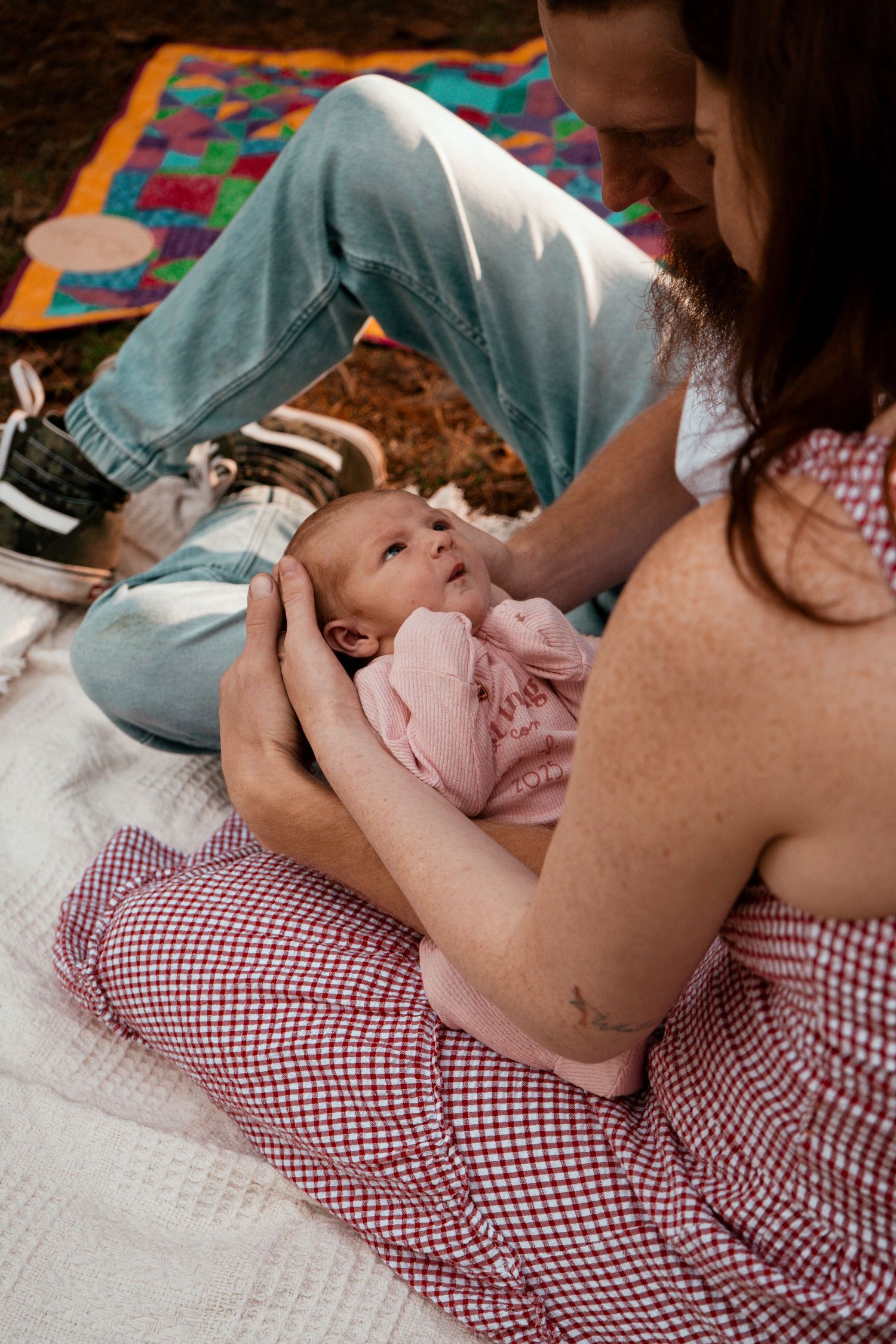 Baby laying on red checkered blanket outdoors with colorful quilt nearby.