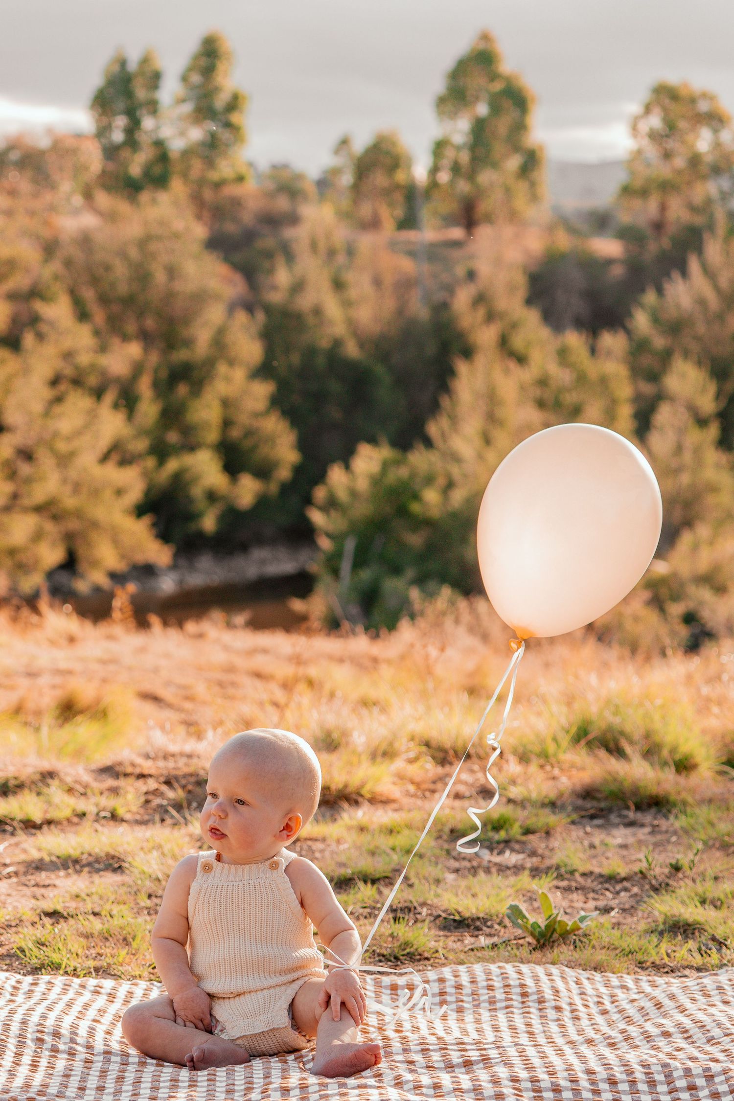 Baby in white sweater sits on picnic blanket with pink balloon during golden hour sunset in autumn.
