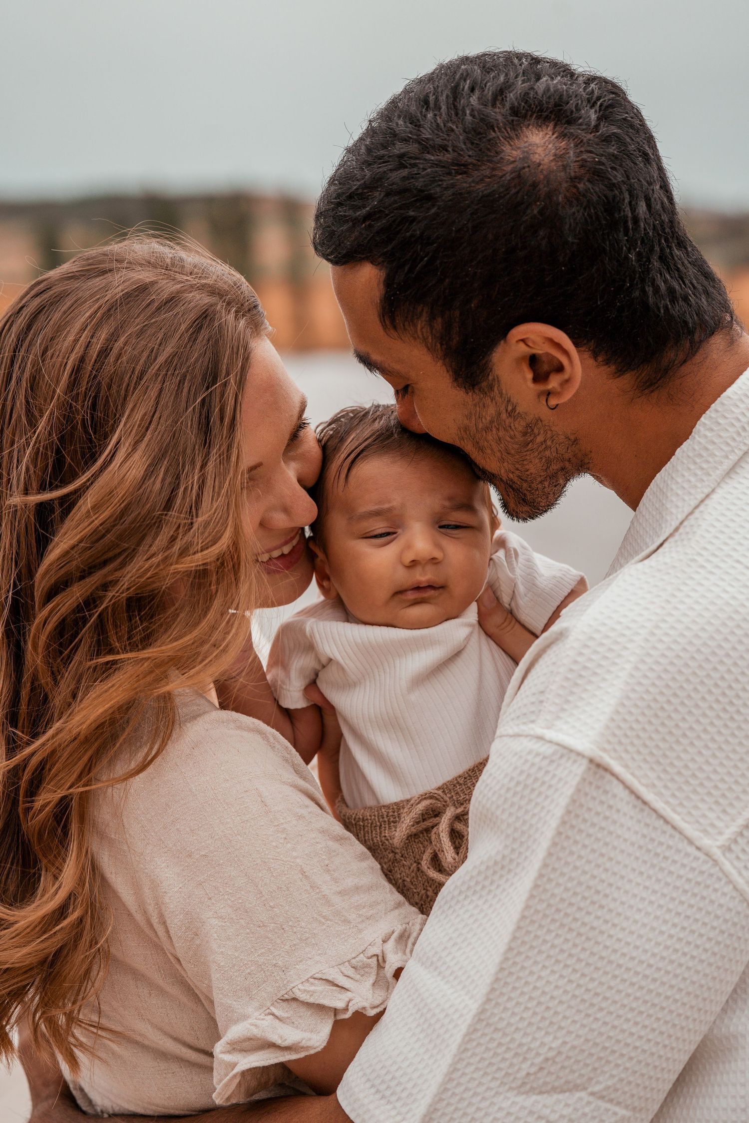A tender family moment as parents share loving kisses with their sleeping baby outdoors at sunset.